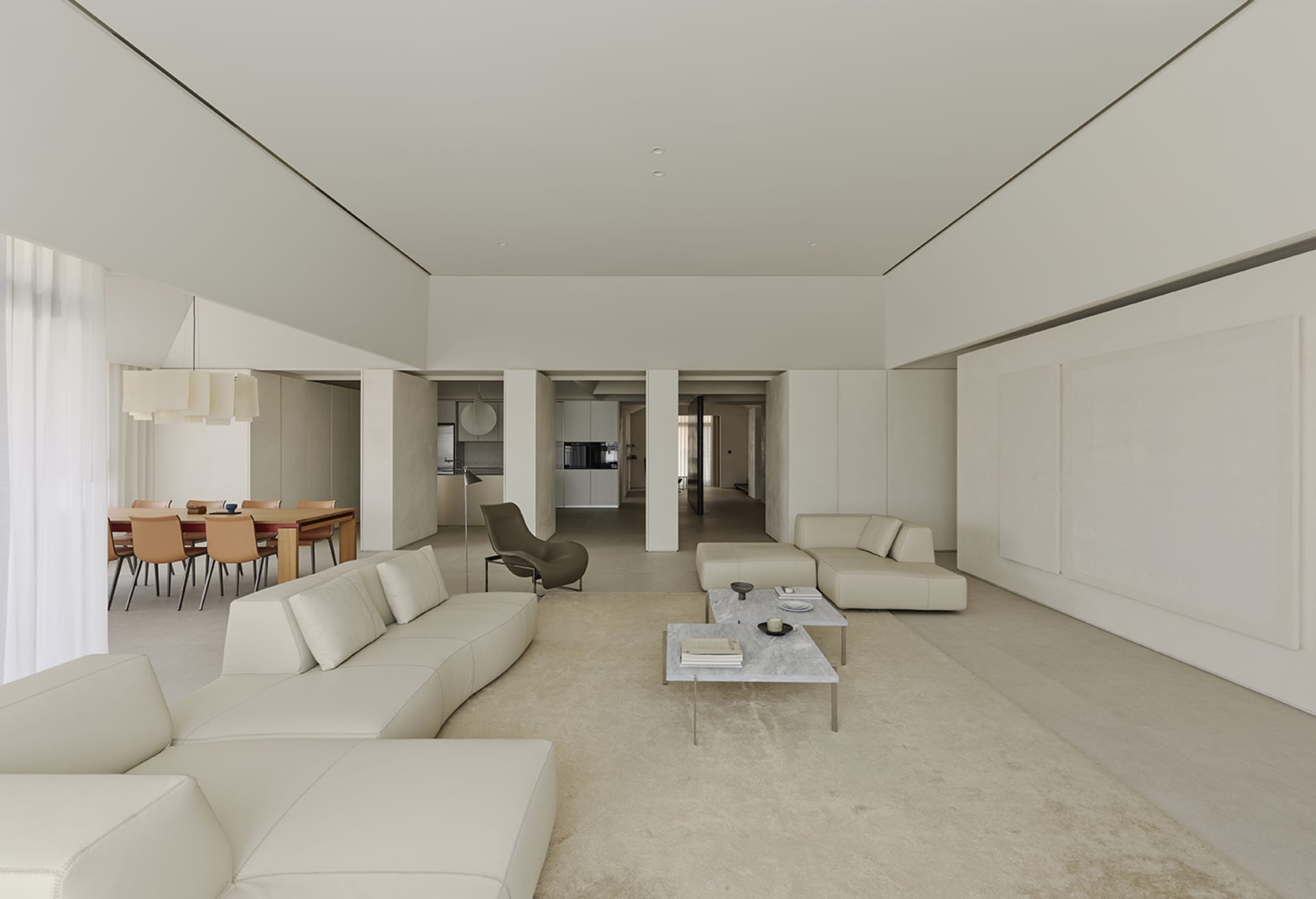 Bright and airy open-plan living and dining area featuring a large curved white leather sofa, marble coffee tables, a sculptural dark green lounge chair, and a wooden dining table with terracotta chairs in the background.