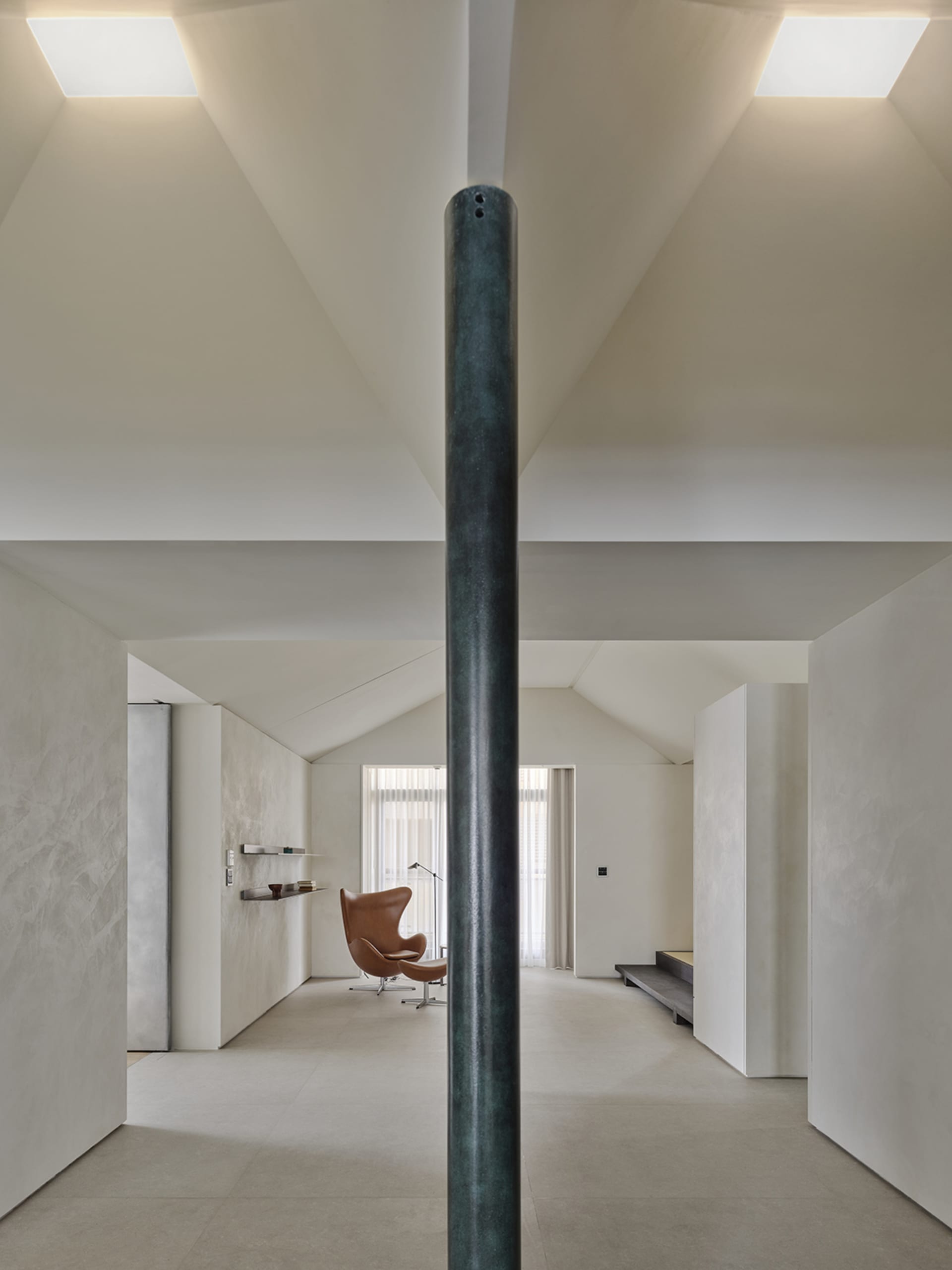 Modern minimalist living room with high sloped concrete ceiling, large aged green steel pillar in foreground, brown leather Egg chair in background, and soft natural light.