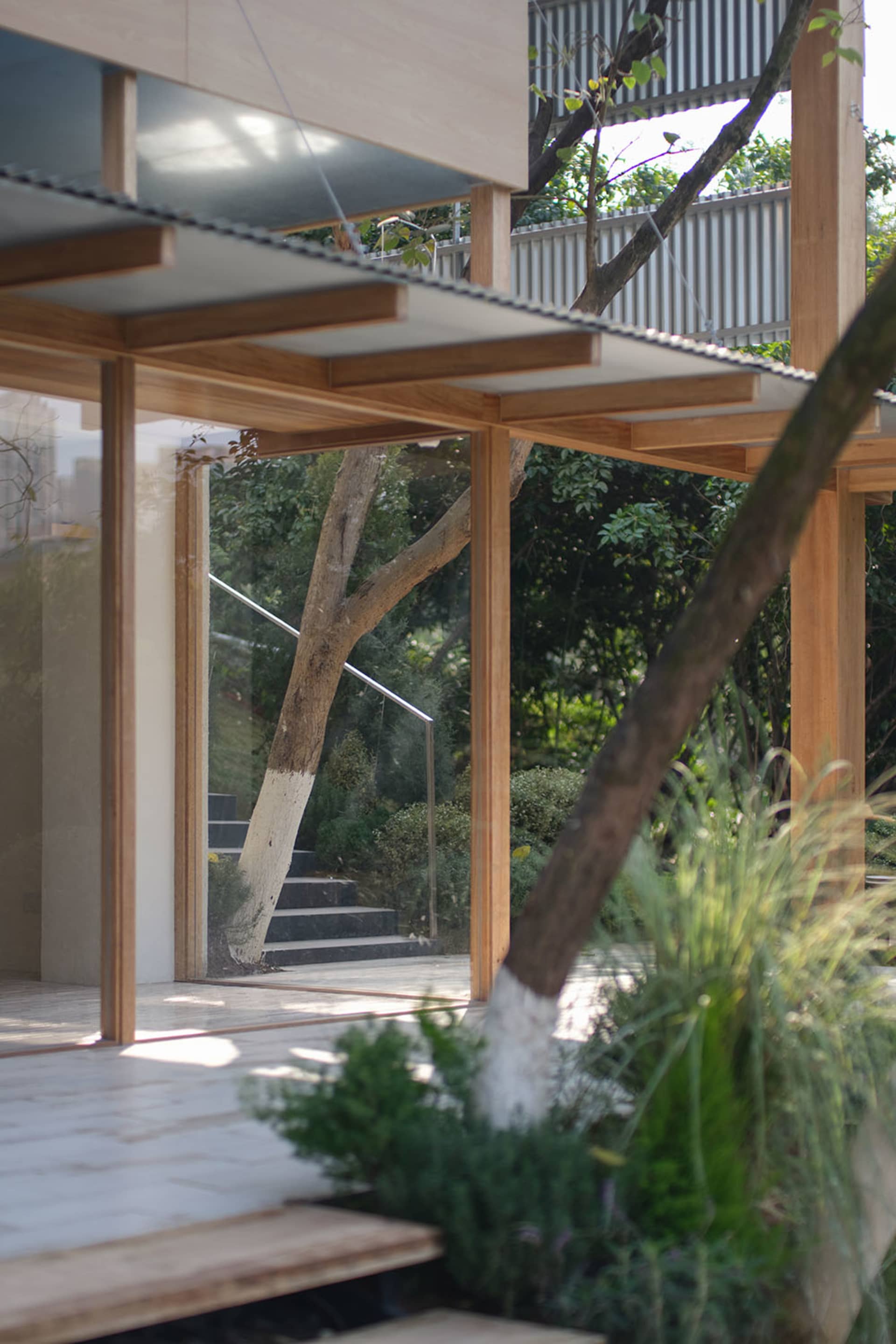 Modern house interior featuring warm teak wood beams and large glass openings that frame a lush tropical garden with trees and stone steps.