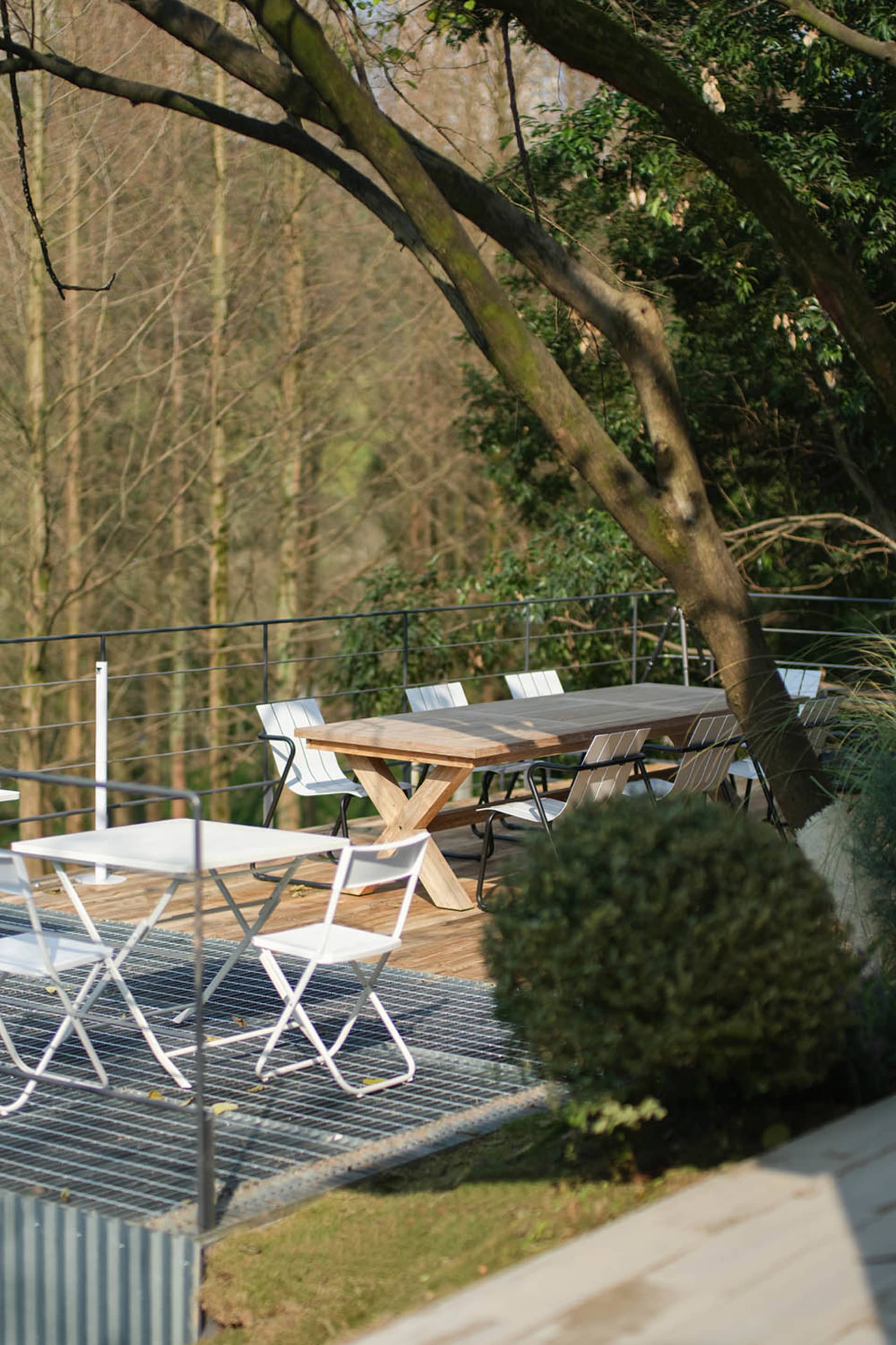 Elevated wooden deck with teak dining table, white folding chairs, and metal railing overlooking a dense forest canopy.