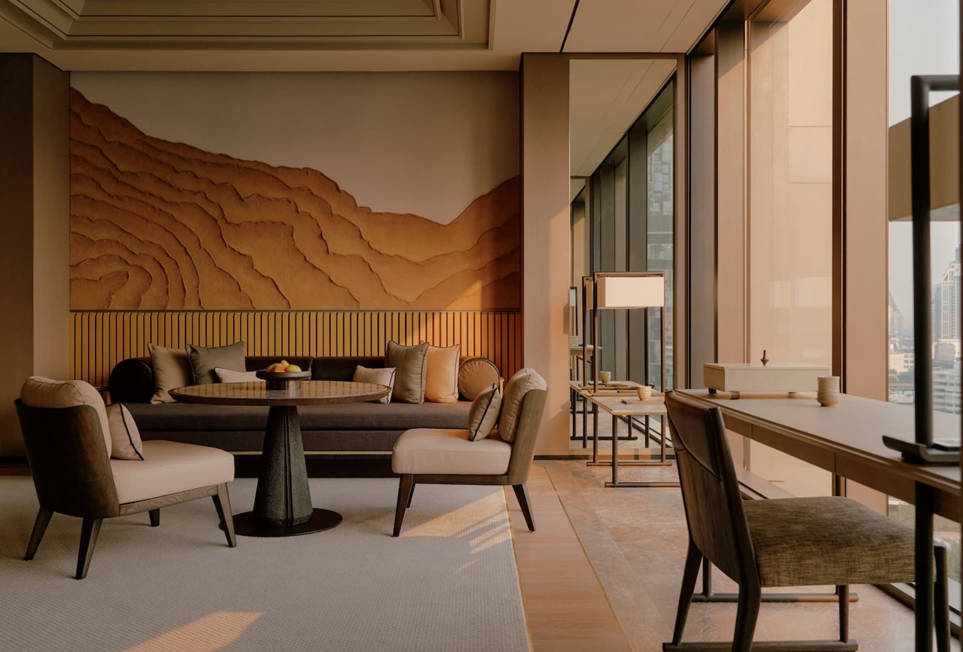 Modern luxury living room with a large terracotta layered mountain mural, walnut wood armchairs, a round pedestal table, and a dark grey sofa with neutral cushions, overlooking a city view through floor-to-ceiling windows.