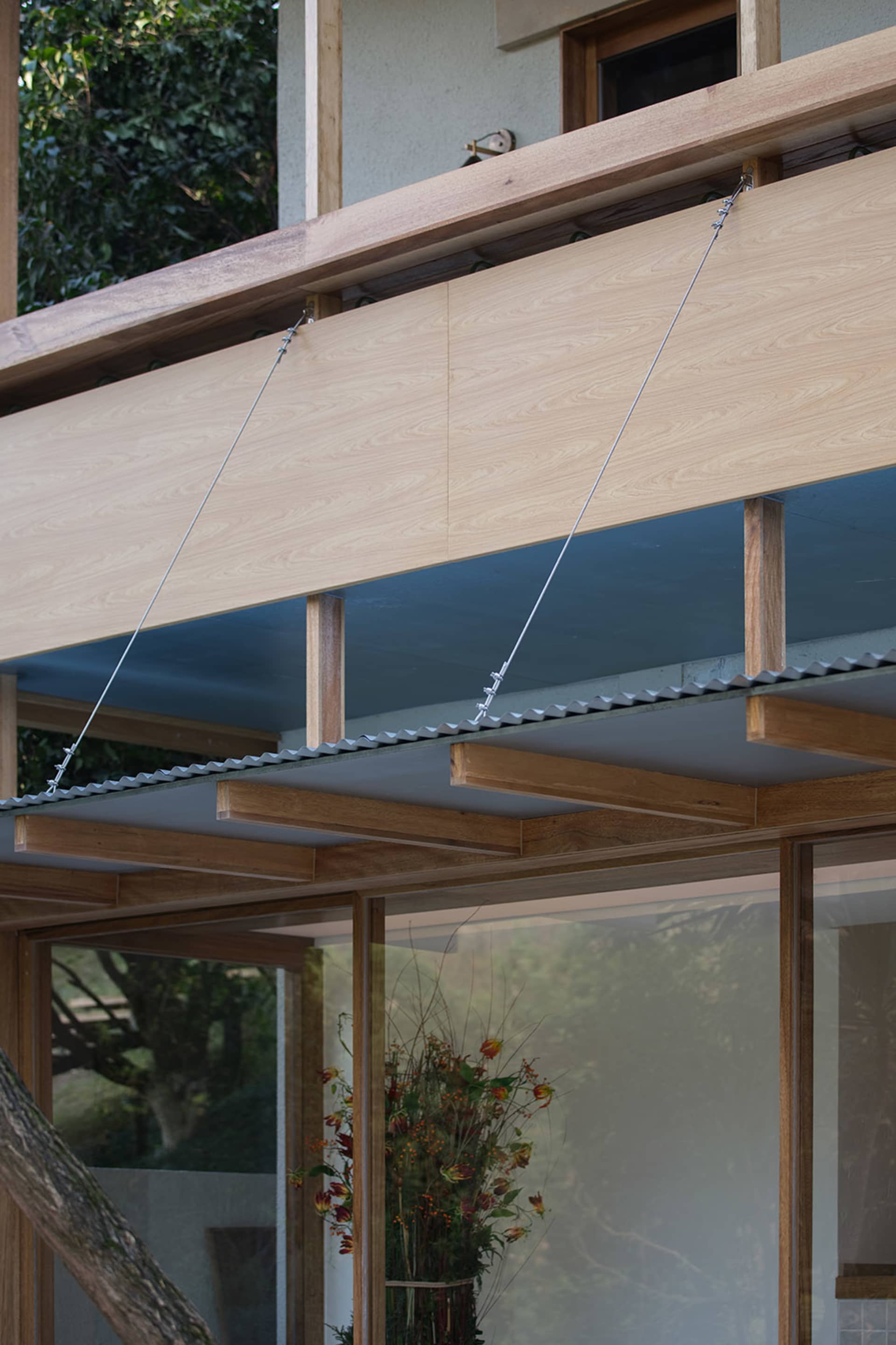 Close-up view of light-colored cypress wooden eaves suspended by steel tension cables, showing structural wooden beams, corrugated metal roofing, and large glass windows below.