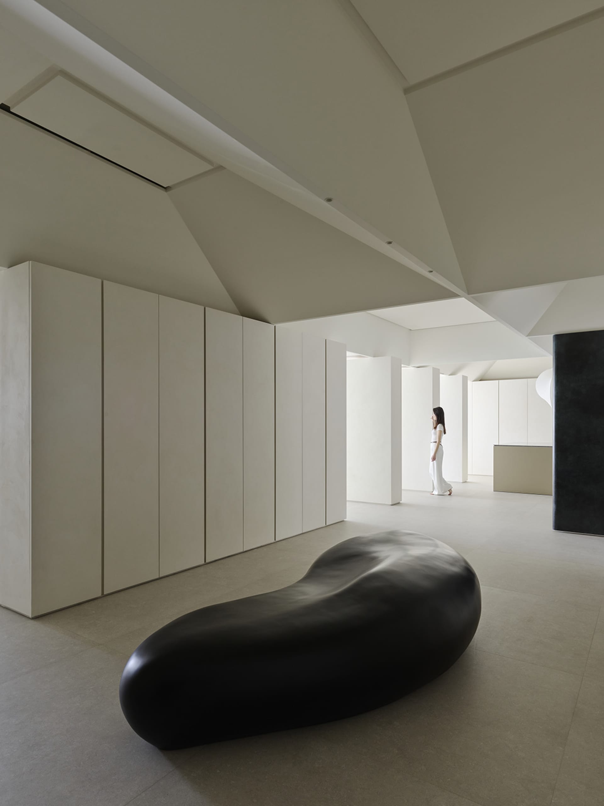 Minimalist gallery interior featuring a large black organic-shaped sculpture in the foreground, floor-to-ceiling micro-cement walls, and a woman in white standing in the distance under dramatic angular ceilings.