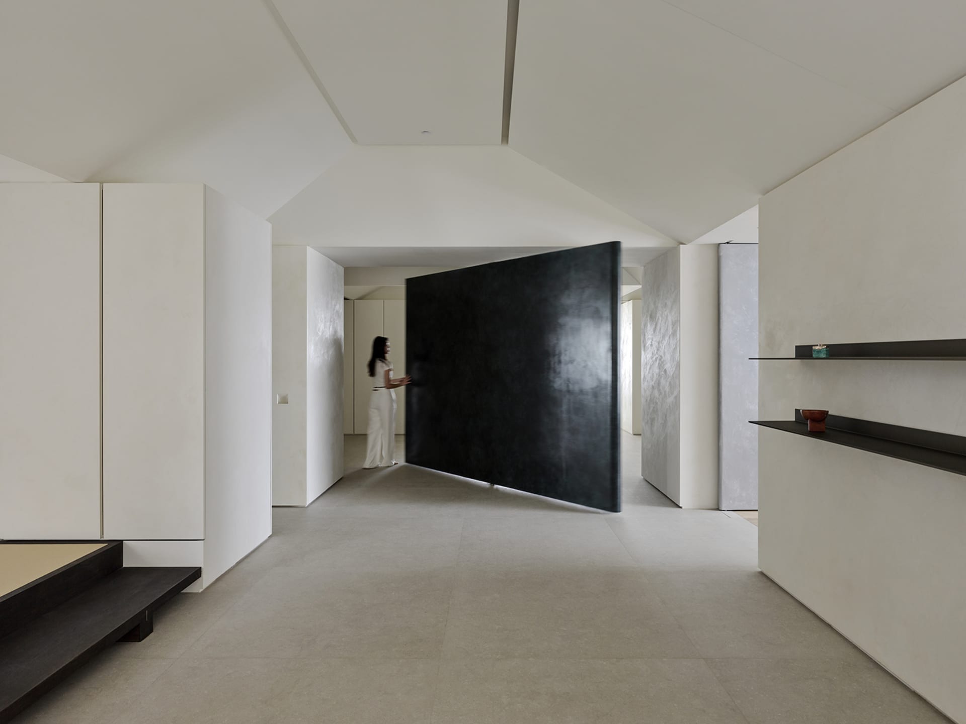 Minimalist Japanese-style entryway featuring a large black pivot door, micro-cement walls and floors, built-in storage, and floating dark shelves with a woman in white standing in the background.