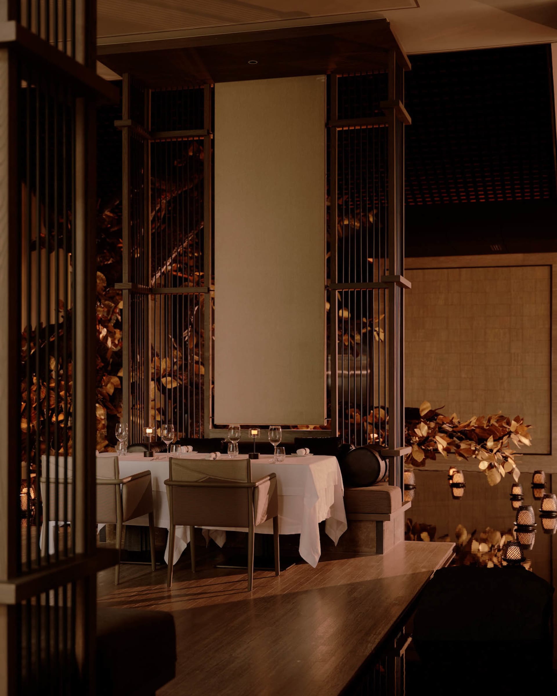 Elegant fine-dining restaurant interior featuring dark wooden lattice room dividers, white tablecloth dining tables, and golden leaf wall art under warm lighting.