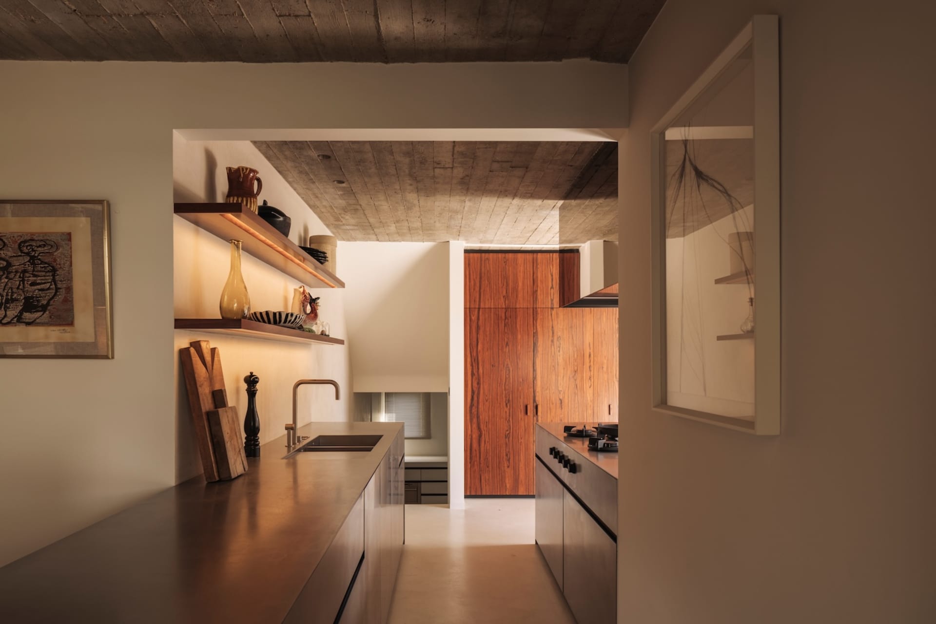 Modern minimalist kitchen featuring warm walnut wood cabinetry, concrete ceiling, stainless steel countertops, and open shelving with curated ceramics.