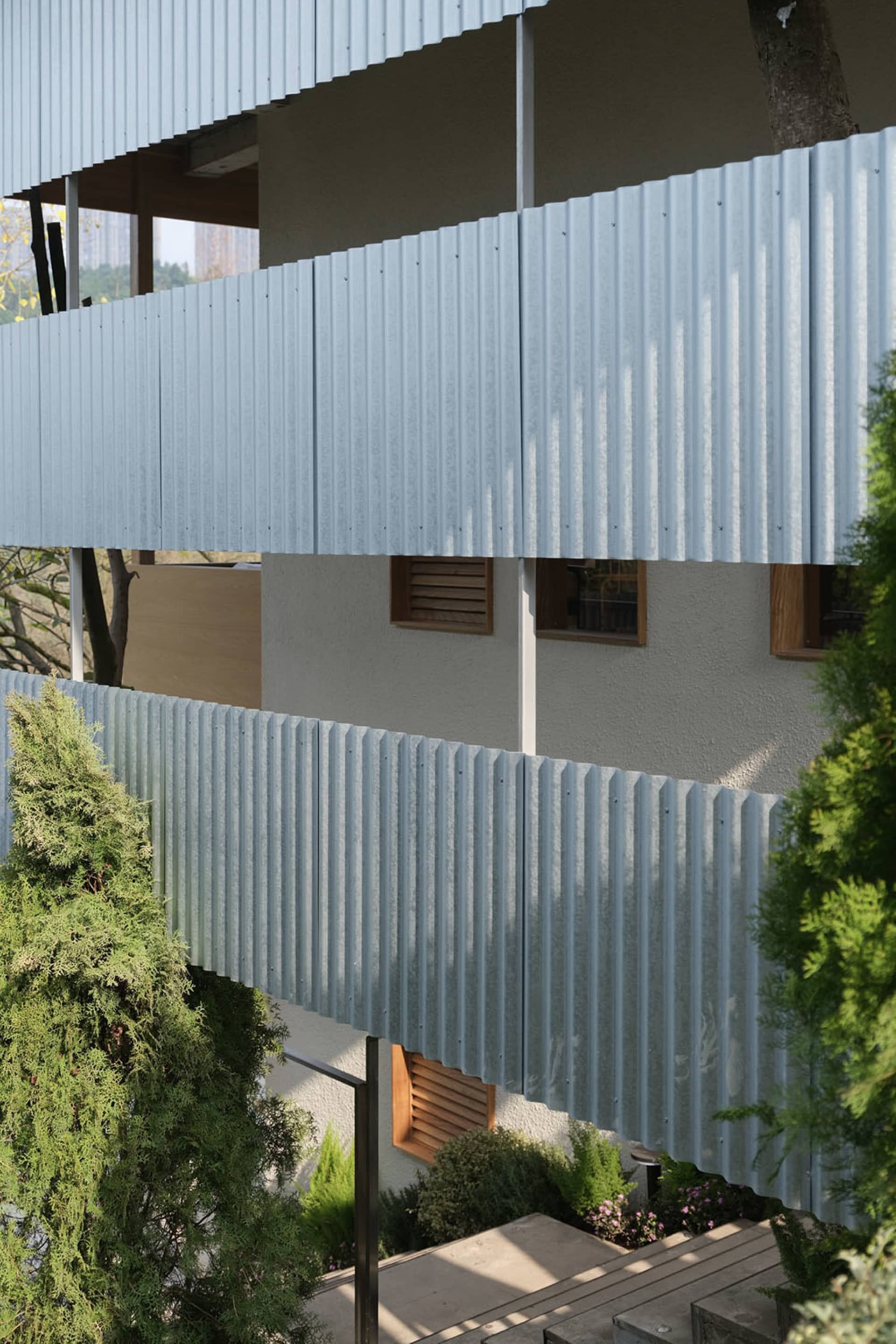 Modern residential building exterior featuring light blue corrugated metal cladding, wooden louvered windows, and surrounding evergreen trees and shrubs.