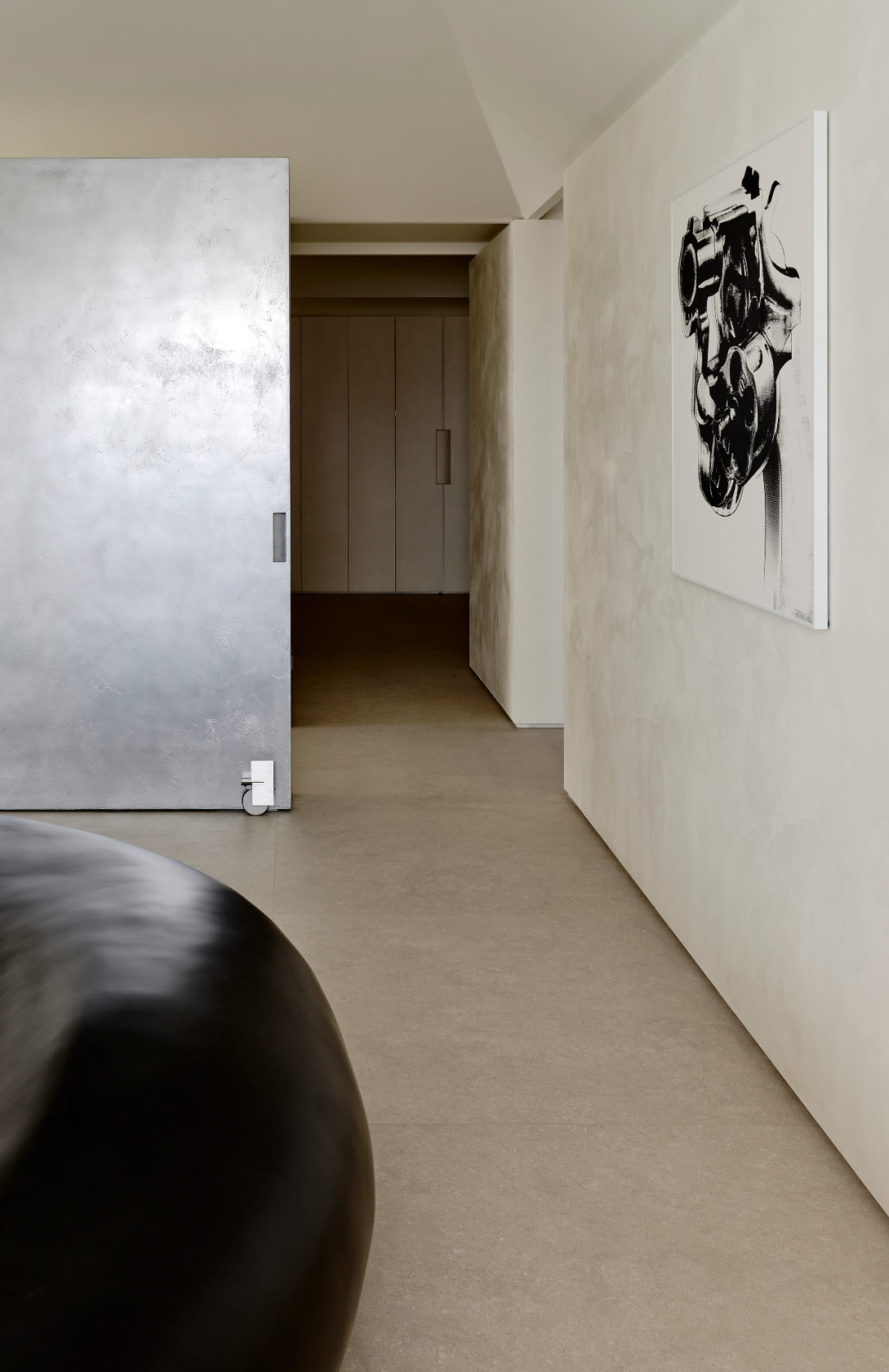 Modern minimalist hallway featuring seamless micro-cement walls, concrete floor, a large black sculptural object in the foreground, and a black and white abstract engine artwork hanging on the right wall.