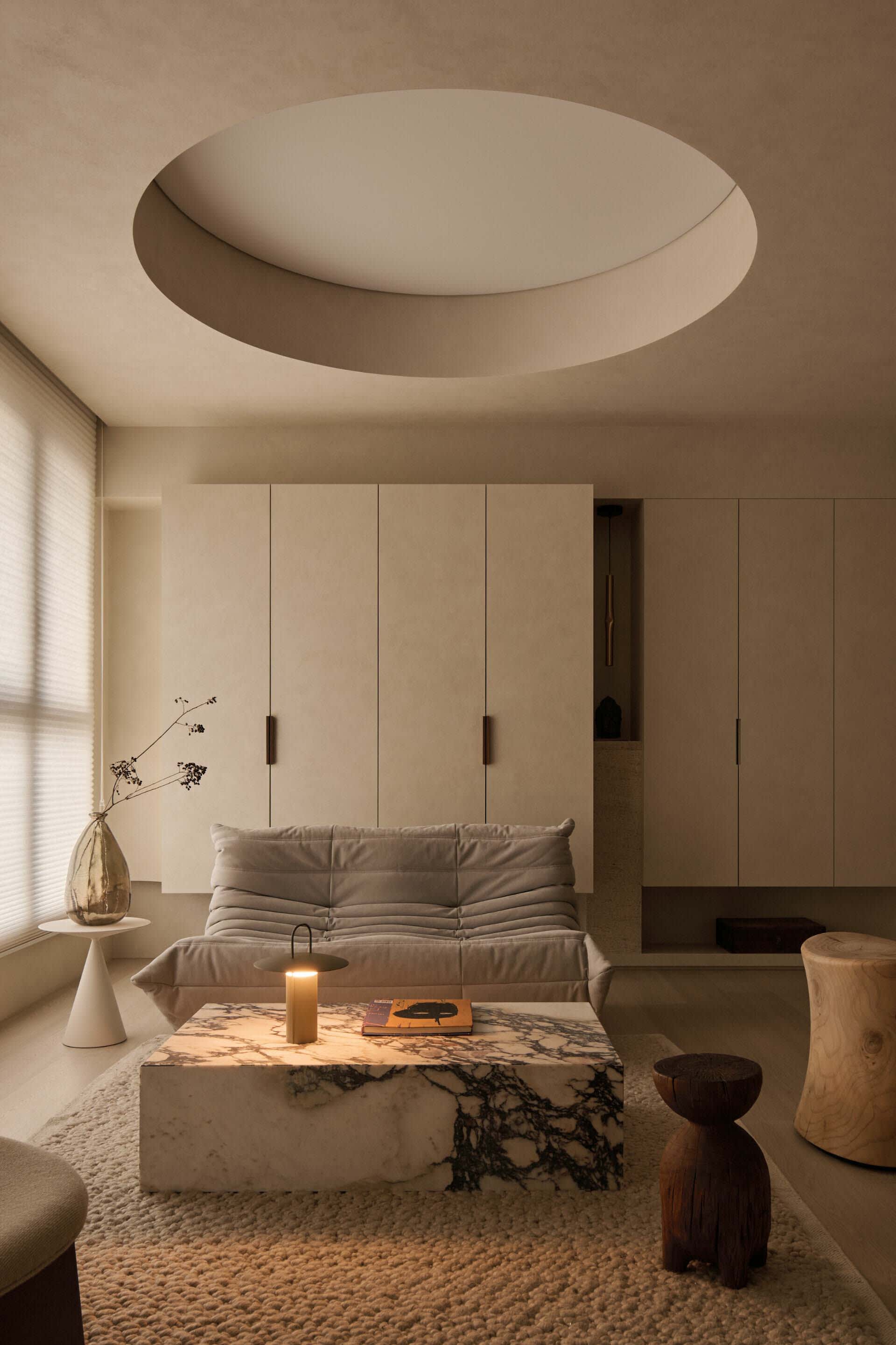 Minimalist living room featuring a large Calacatta marble coffee table, a modular beige sofa, textured boucle rug, and a dramatic circular recessed ceiling light.