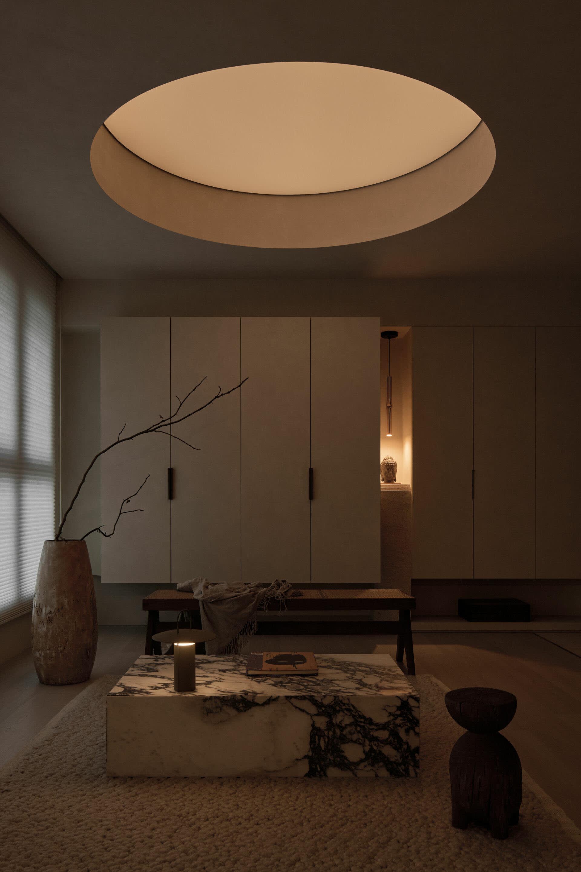 Minimalist living room featuring a large marble coffee table, low wooden bench, textured beige walls, and a dramatic circular ceiling light fixture.