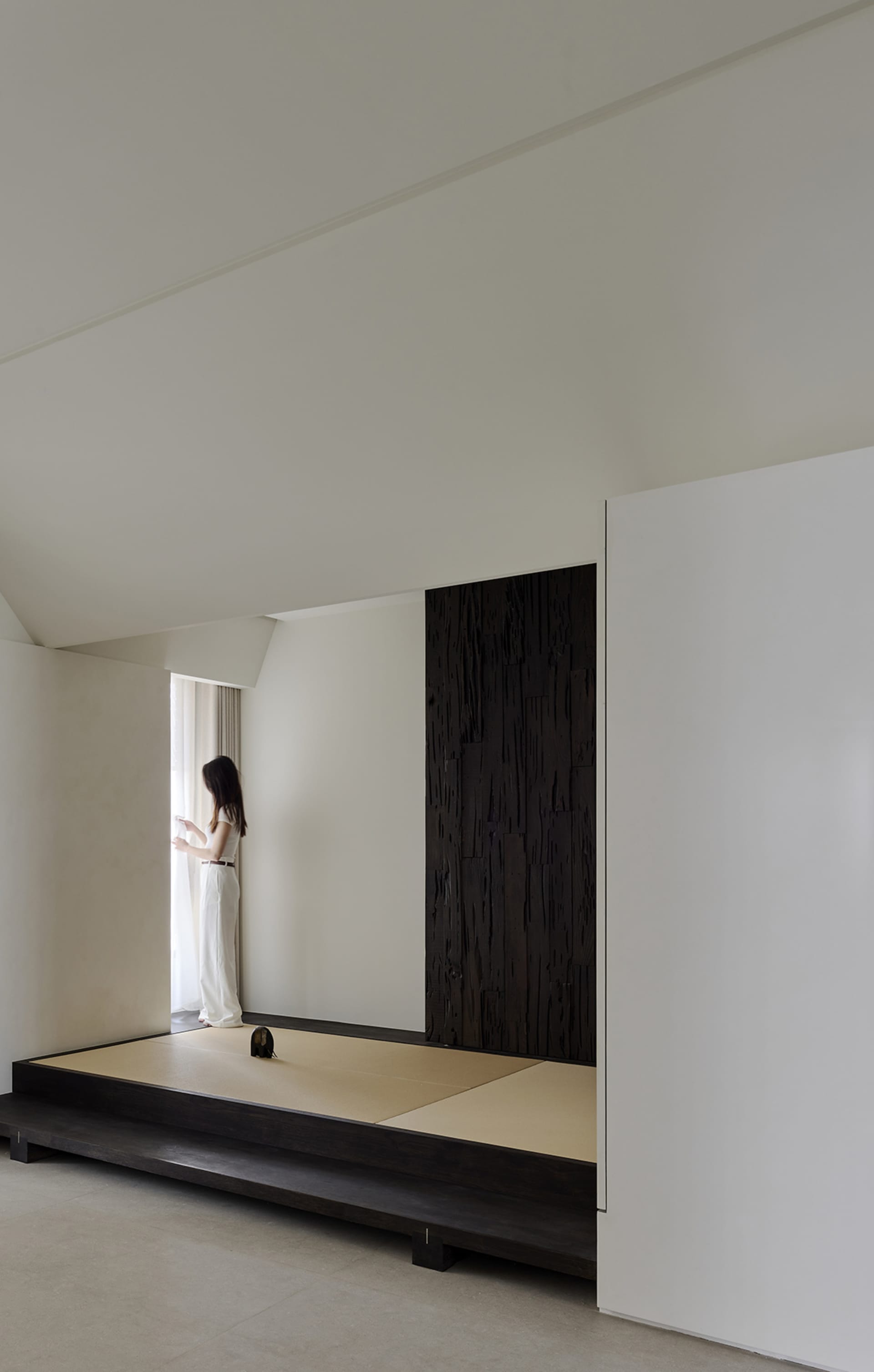 Woman standing by window in a minimalist Japanese-style tatami room featuring a dramatic charred wood wall and raised platform.