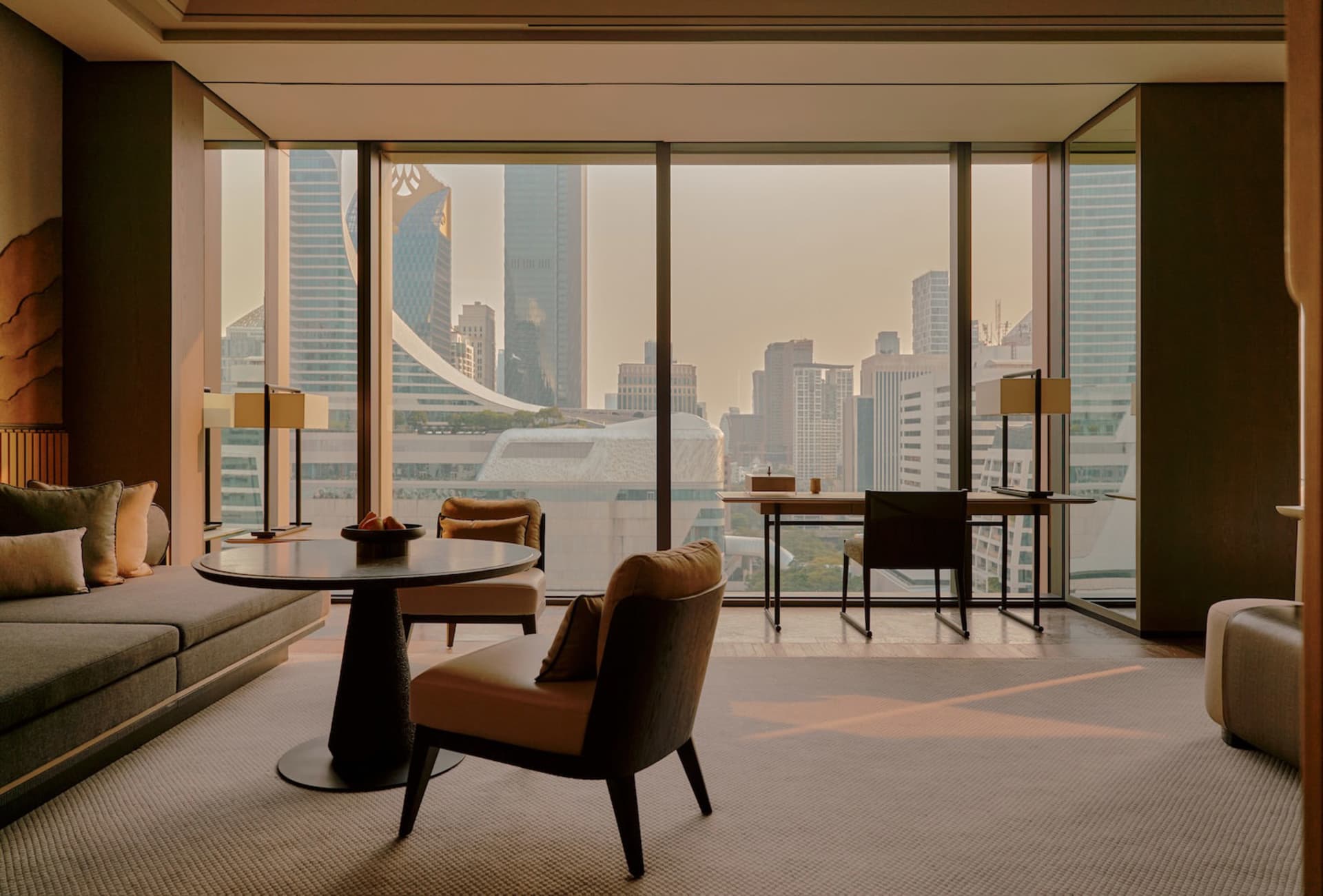 Modern luxury living room with curved walnut table, taupe leather armchairs, built-in sofa, and dramatic floor-to-ceiling windows overlooking the Hong Kong skyline at dusk.