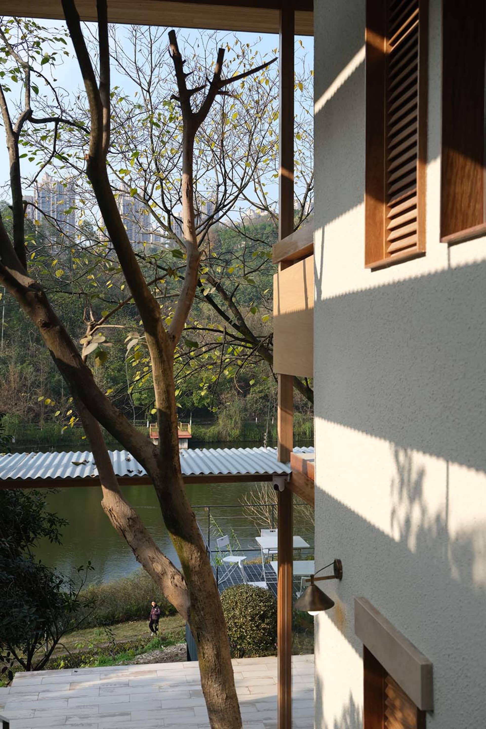 View from a modern house terrace featuring wooden window frames and shutters, overlooking a lake with trees and outdoor seating area below.
