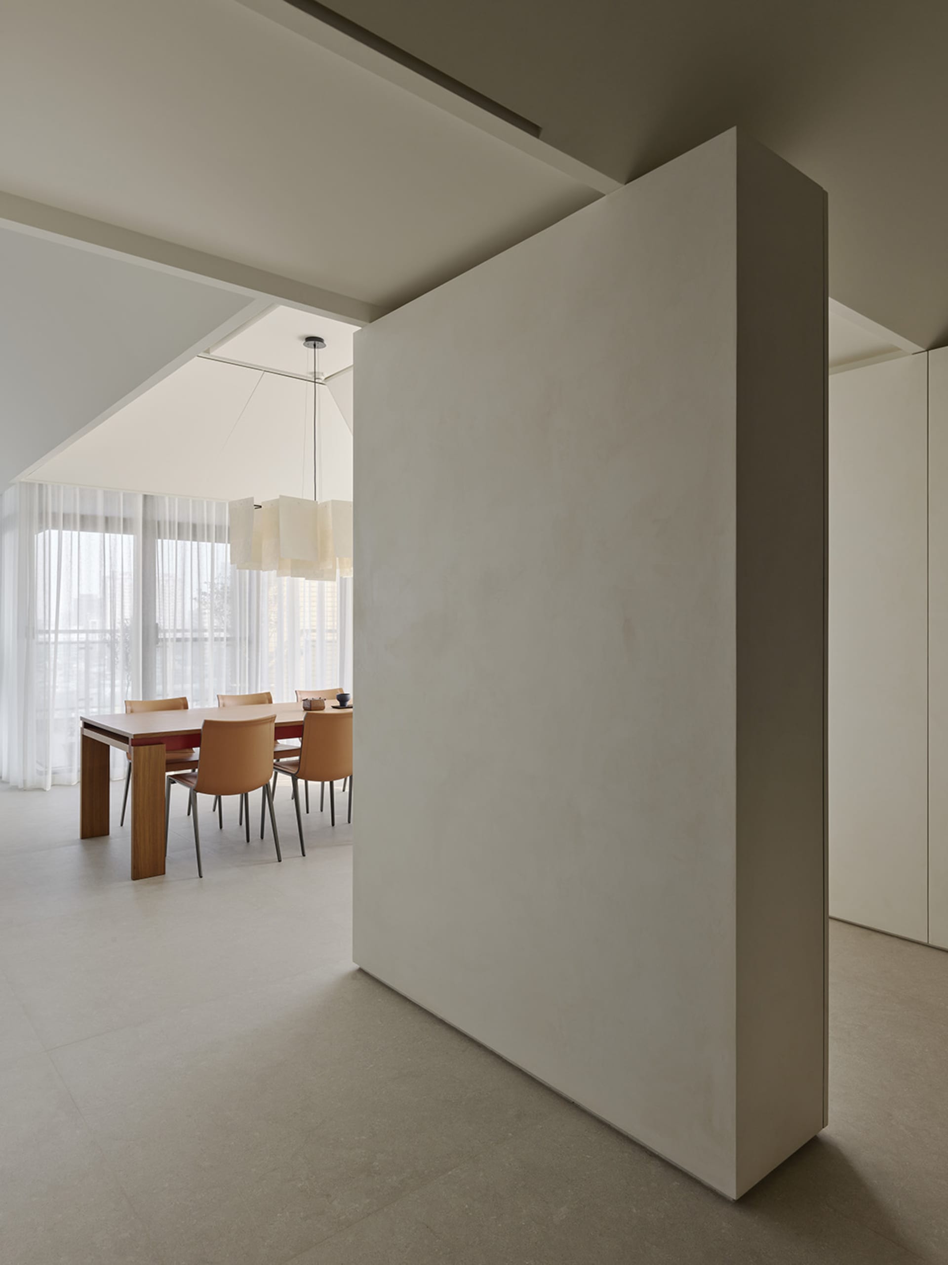 Modern minimalist dining room featuring a large micro-cement partition wall, wooden dining table, tan leather chairs, and a sculptural pendant light under a high sloped ceiling.