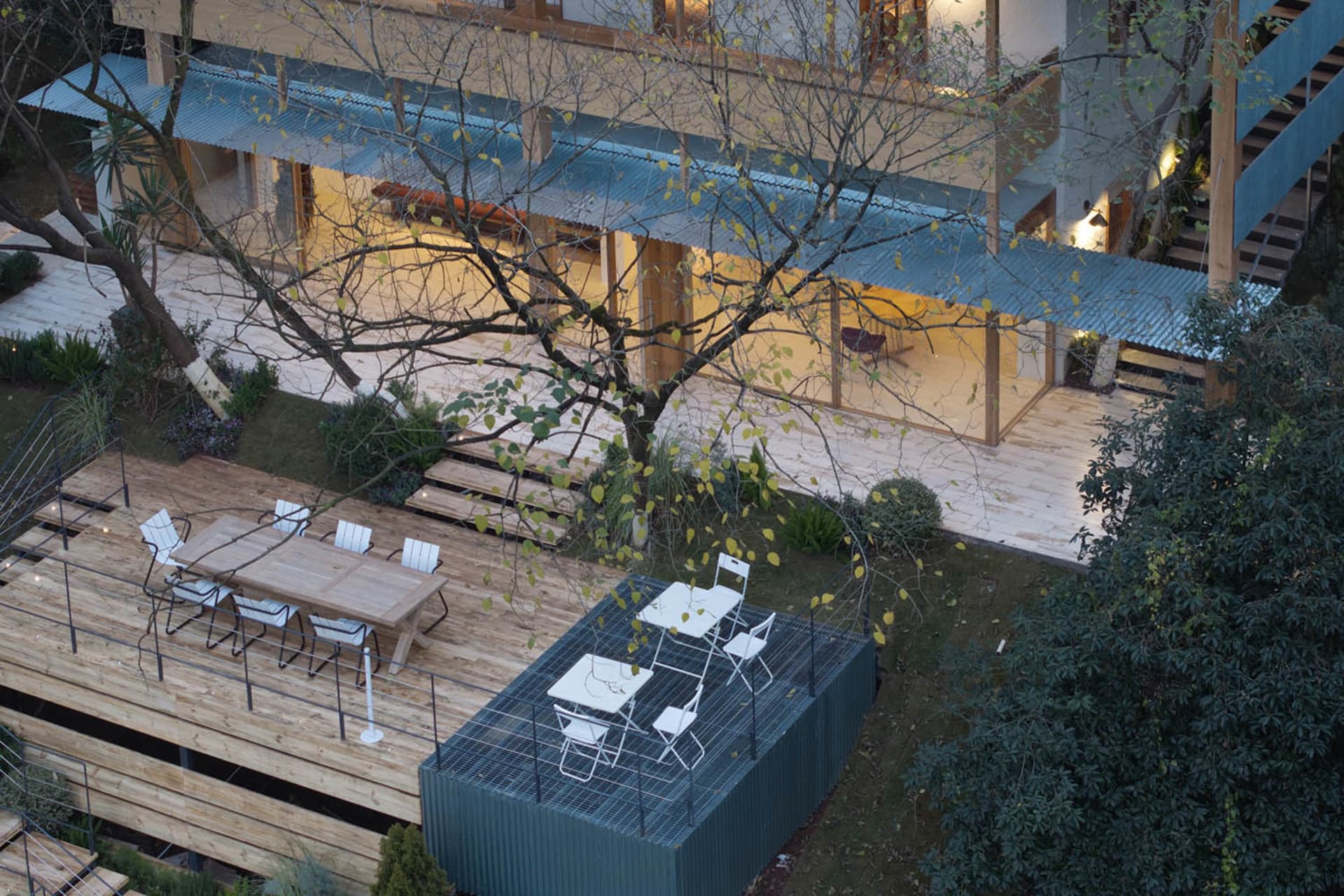 Aerial view of a modern house backyard featuring two distinct outdoor dining areas: a large wooden deck with a long timber table and white chairs, and an elevated Corten steel mesh platform with smaller white dining sets, surrounded by trees and integrated with the house architecture.