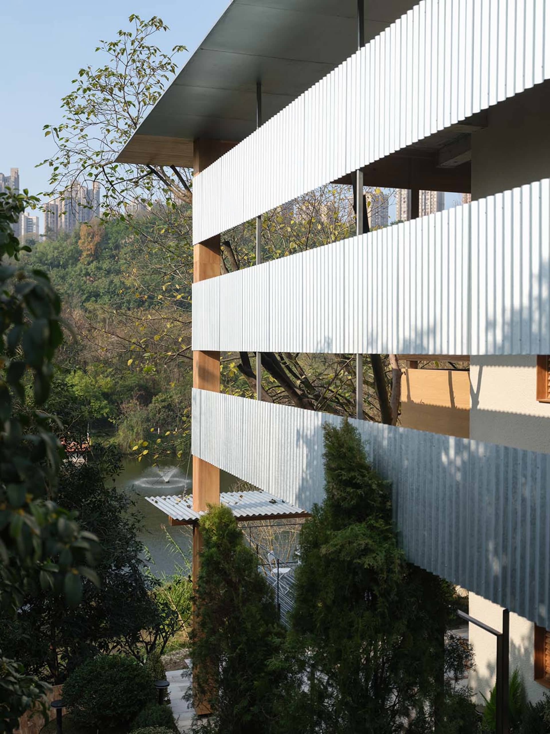 現代山坡住宅建築,外牆使用白色波浪鐵皮、溫潤木柱與大挑簷,俯瞰設有噴泉的庭園水池,周圍環繞茂密綠植。