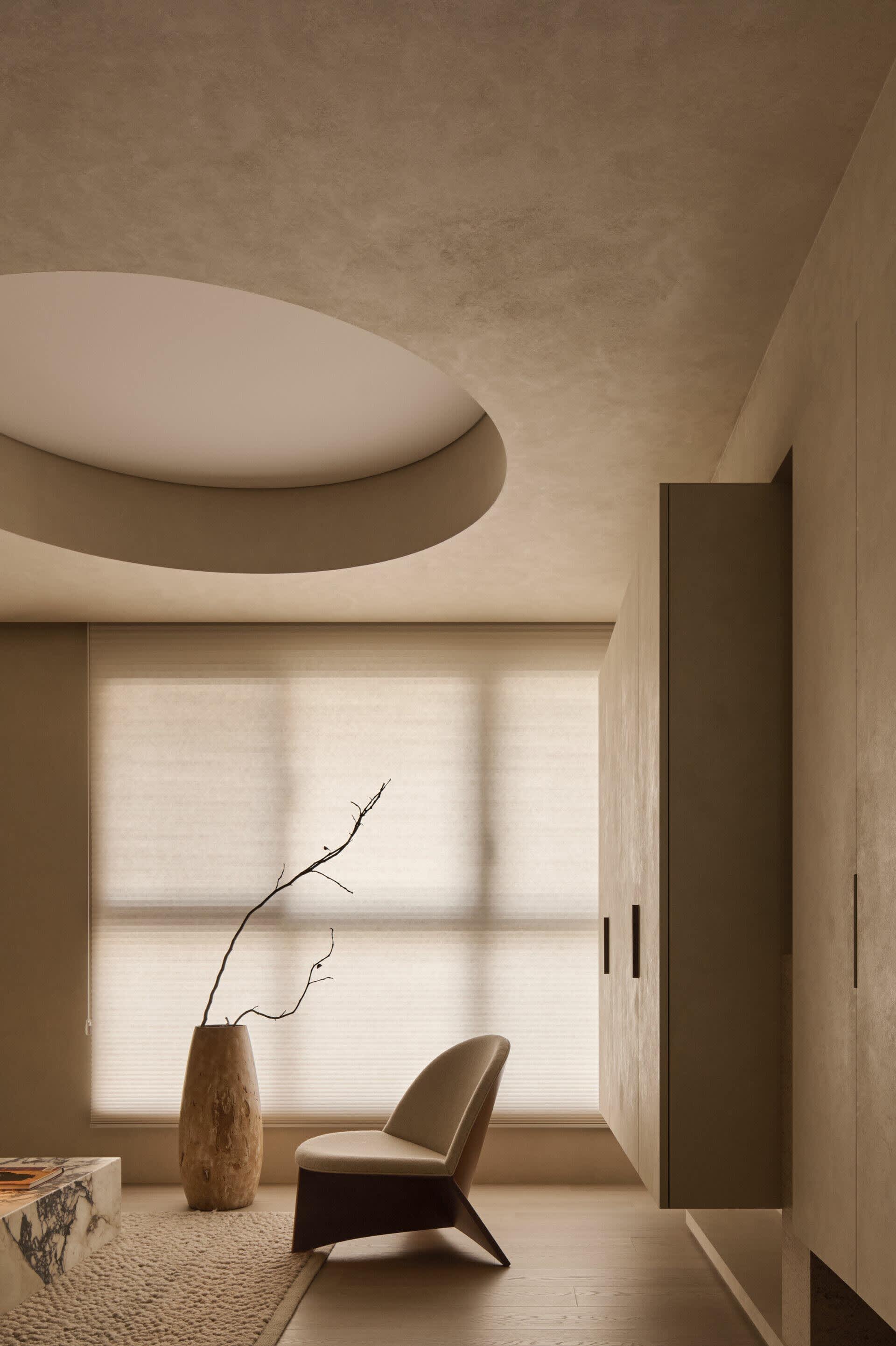 Minimalist living room interior with beige micro-cement walls, curved ceiling feature, beige upholstered reading chair, wooden vase with dried branch, marble coffee table, and textured area rug.