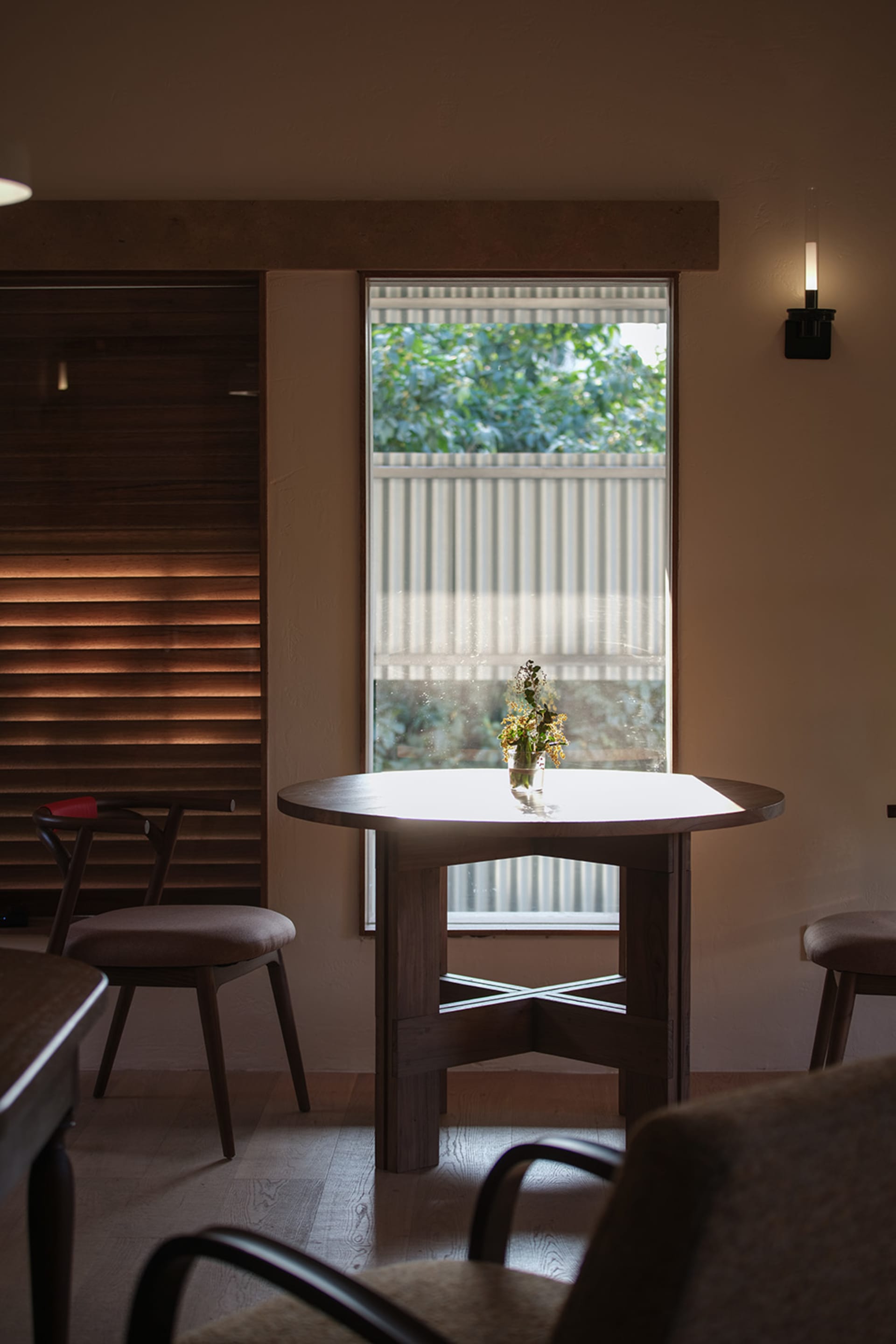 Modern minimalist dining area featuring a solid walnut round table, matching wooden chairs with upholstered seats, warm wood slat wall, and a large window with garden view.