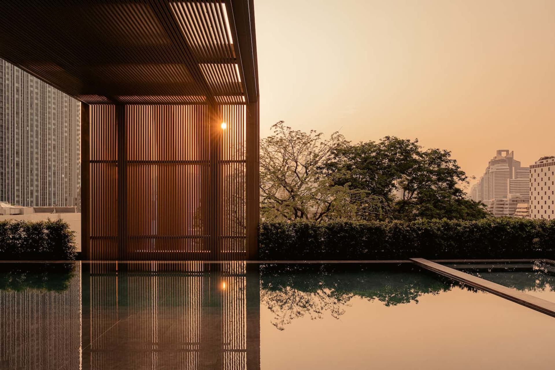 Modern rooftop infinity pool reflecting a dramatic orange sunset, framed by vertical wooden slat screens and a city skyline in the background.