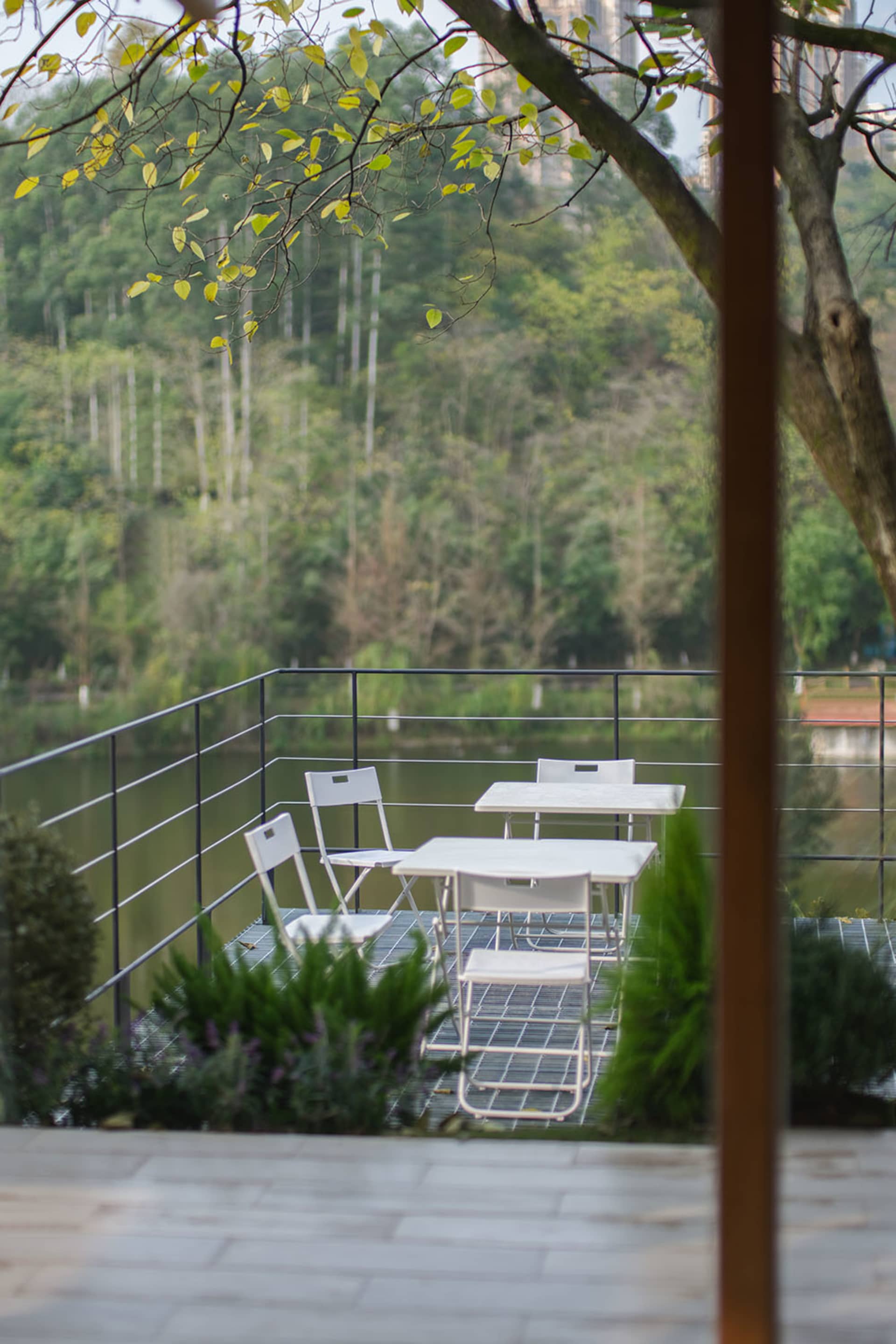White metal dining table and chairs on a wire-mesh terrace overlooking a lake with forest and mountains in the background.