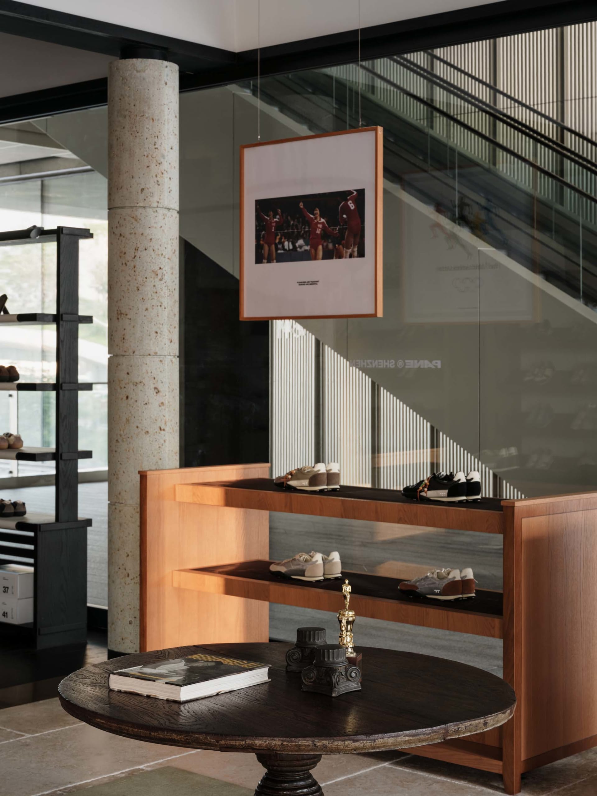 Modern retail interior featuring a large terrazzo column, warm oak floating shelves displaying various sneakers, a round dark wood table with an Oscar statue and book in the foreground, and a framed sports photograph hanging above.
