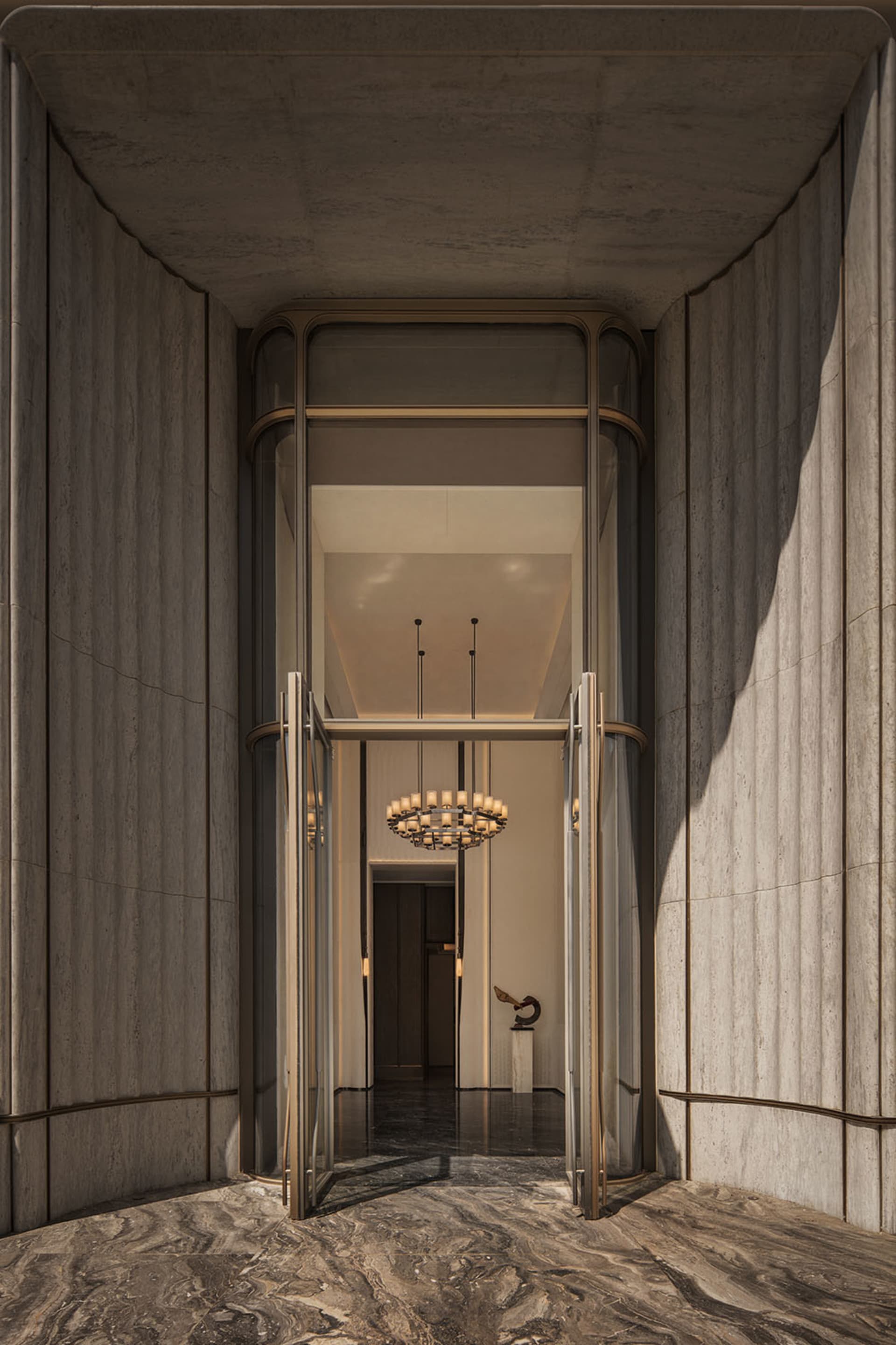 View from inside a luxurious building entrance looking through open ornate brass and glass doors into a grand lobby with a large chandelier, dark marble floor and travertine walls.