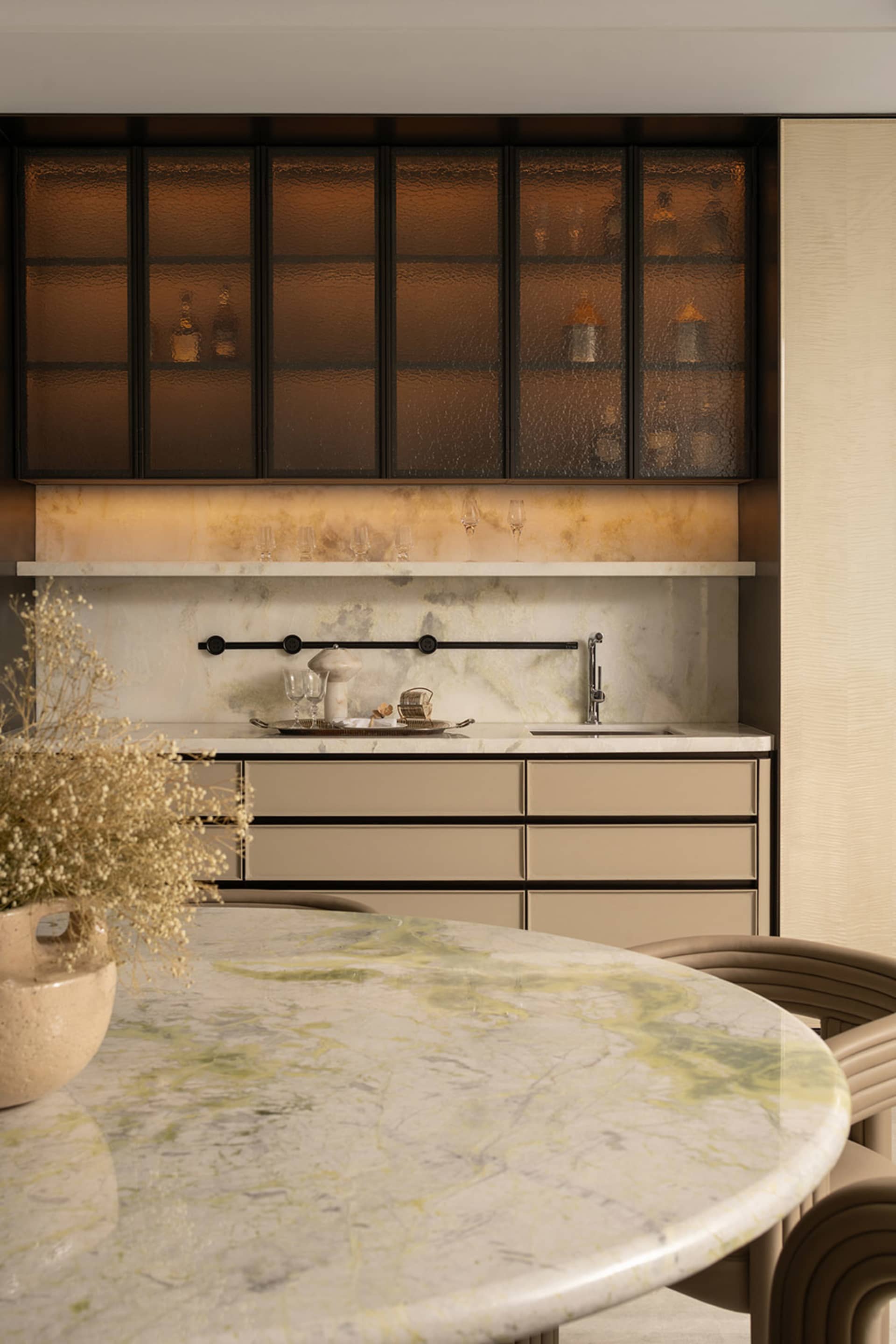 Modern dining room with round Calacatta green marble table, beige curved dining chairs, and a luxurious marble wet bar with amber textured glass upper cabinets and under-cabinet lighting.