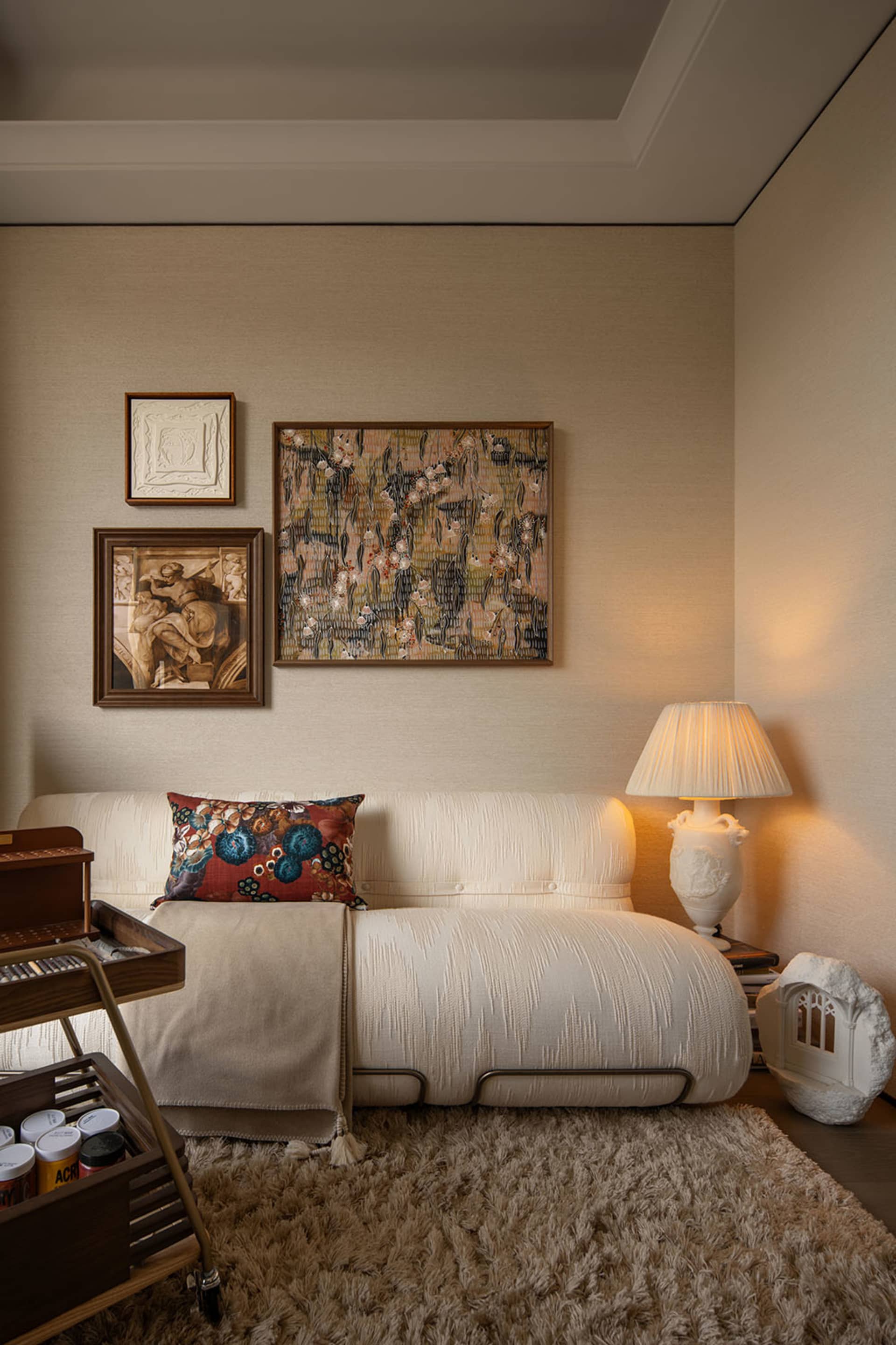 Cream-colored textured sofa with floral pillow, layered gallery wall art, table lamp, and shaggy beige rug in a warm living room corner