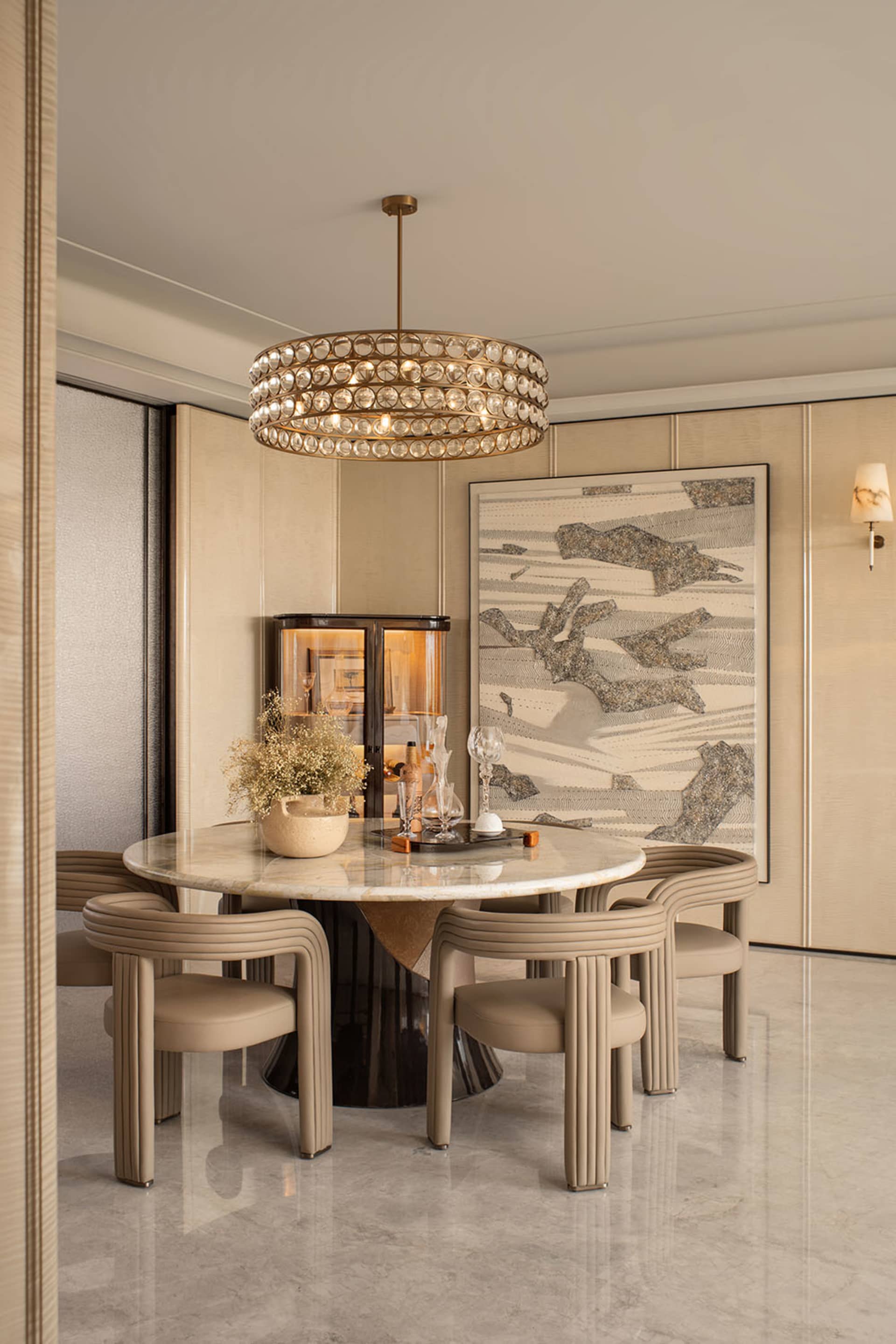 Elegant contemporary dining room with a round Calacatta marble table, fluted beige leather chairs, a large crystal chandelier, and abstract wall art.