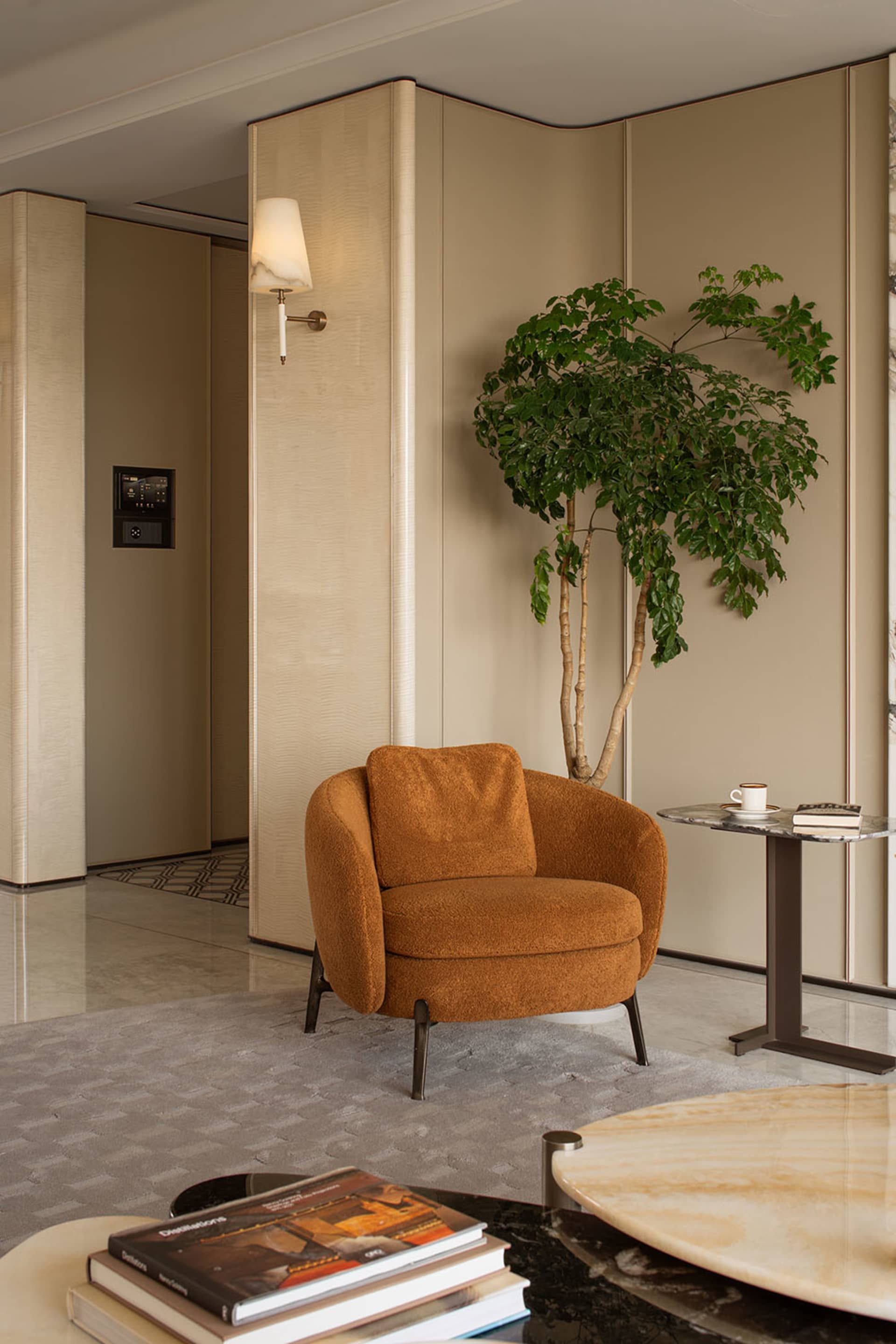 Modern living room corner featuring a burnt orange boucle armchair, tall potted tree, marble side table, and textured beige walls with wall sconce.