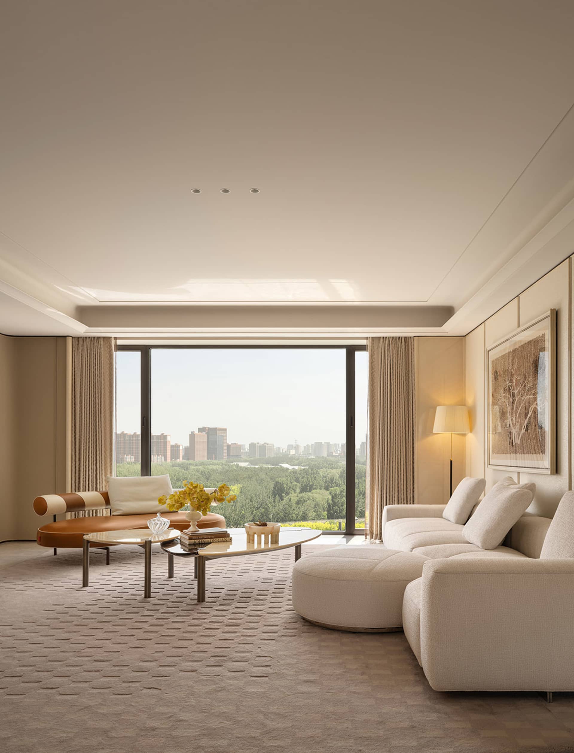 Modern luxury living room featuring a large beige boucle sectional sofa, curved brown and cream leather chaise lounge, glass and metal coffee table, and floor-to-ceiling windows overlooking a green forest and city skyline.