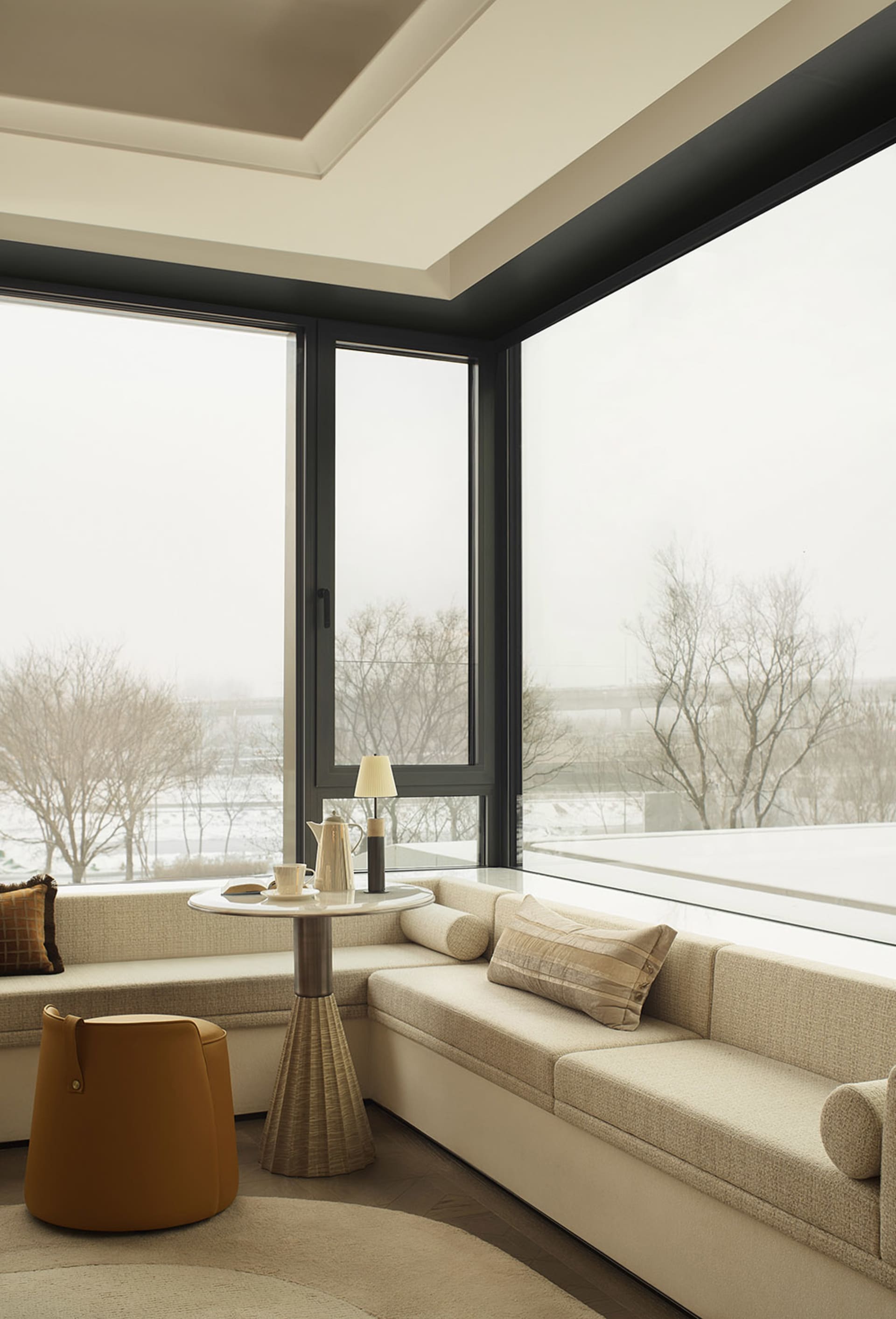 Modern minimalist living room corner featuring a built-in boucle fabric window seat banquette, round fluted side table, mustard yellow leather ottoman, and large black-framed windows with winter view
