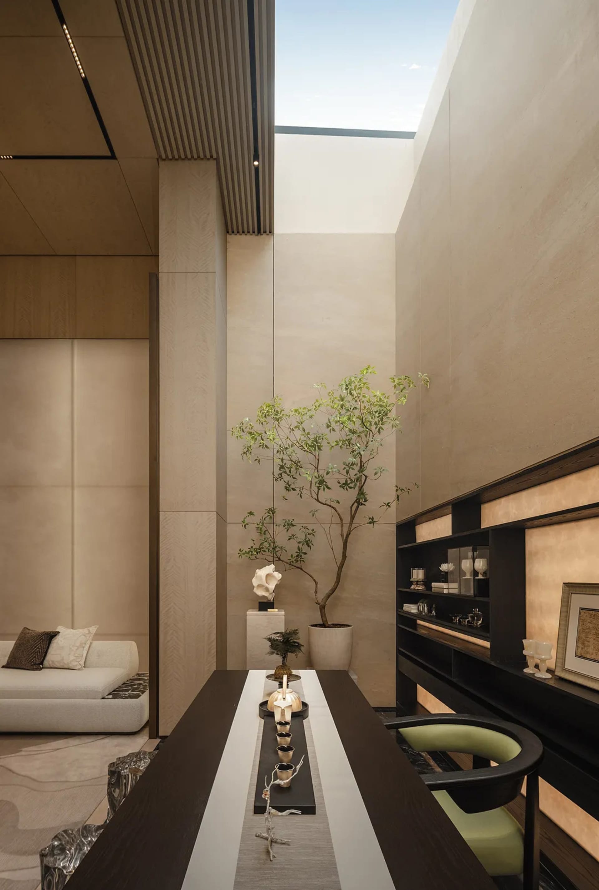 Modern minimalist dining room featuring travertine walls, a long dark wood table, built-in dark shelving, and a tall potted tree under a large skylight.