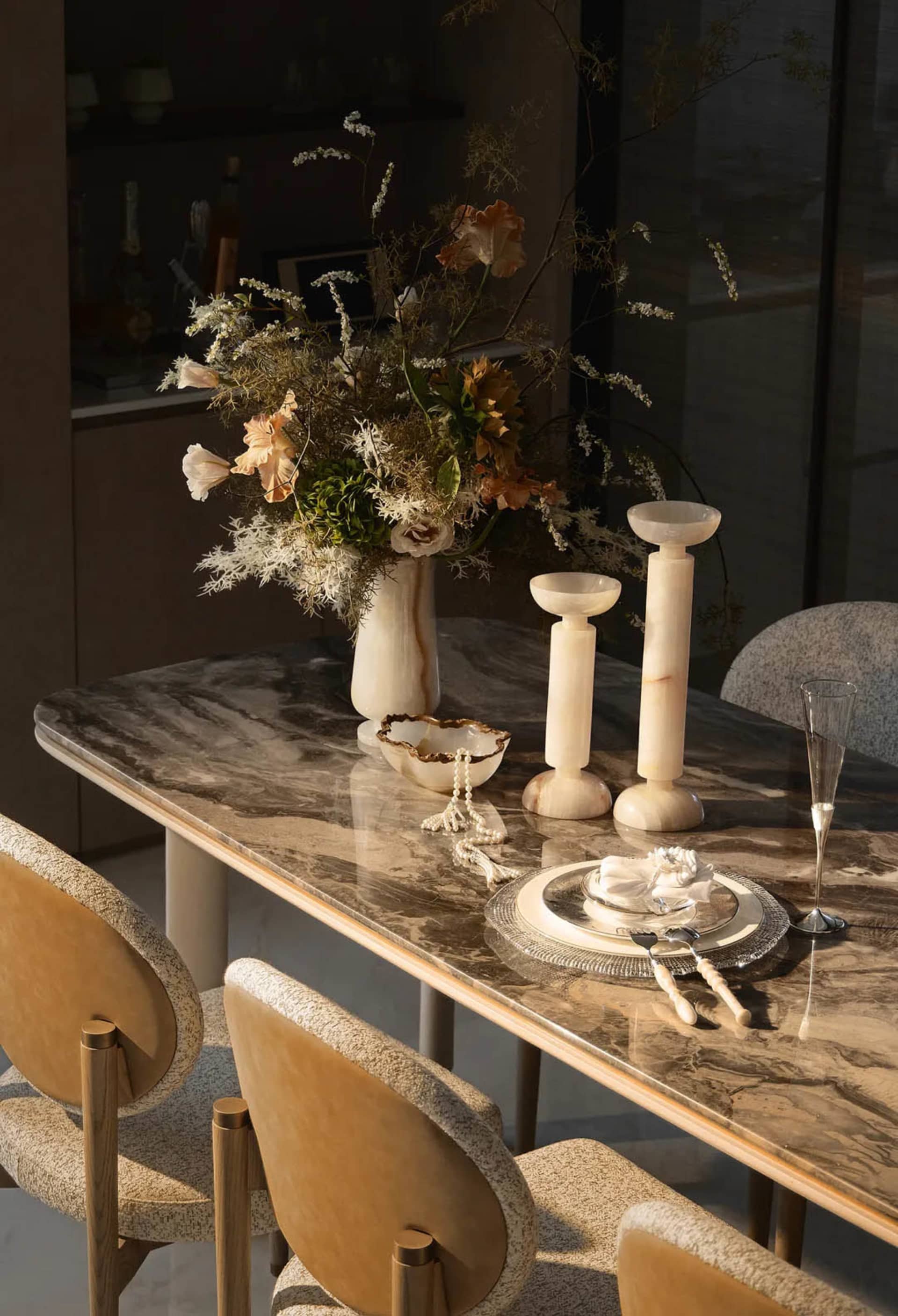 Elegant dining table with brown-veined Calacatta marble top, bouclé upholstered dining chairs, alabaster candle holders, and a large dried floral arrangement in a marble vase.