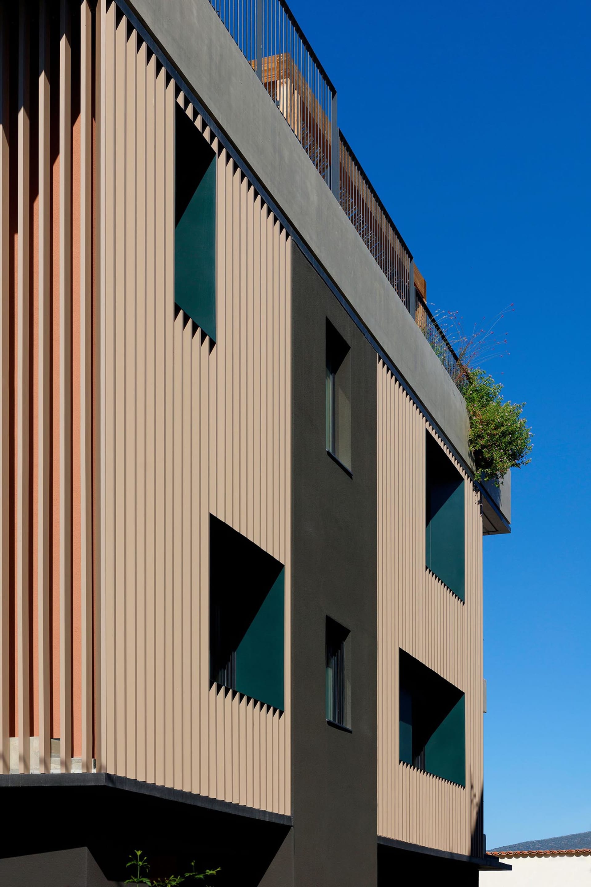 Close-up view of a modern building facade featuring beige corrugated metal panels, dark grey sections, and distinctive triangular windows with green glass.