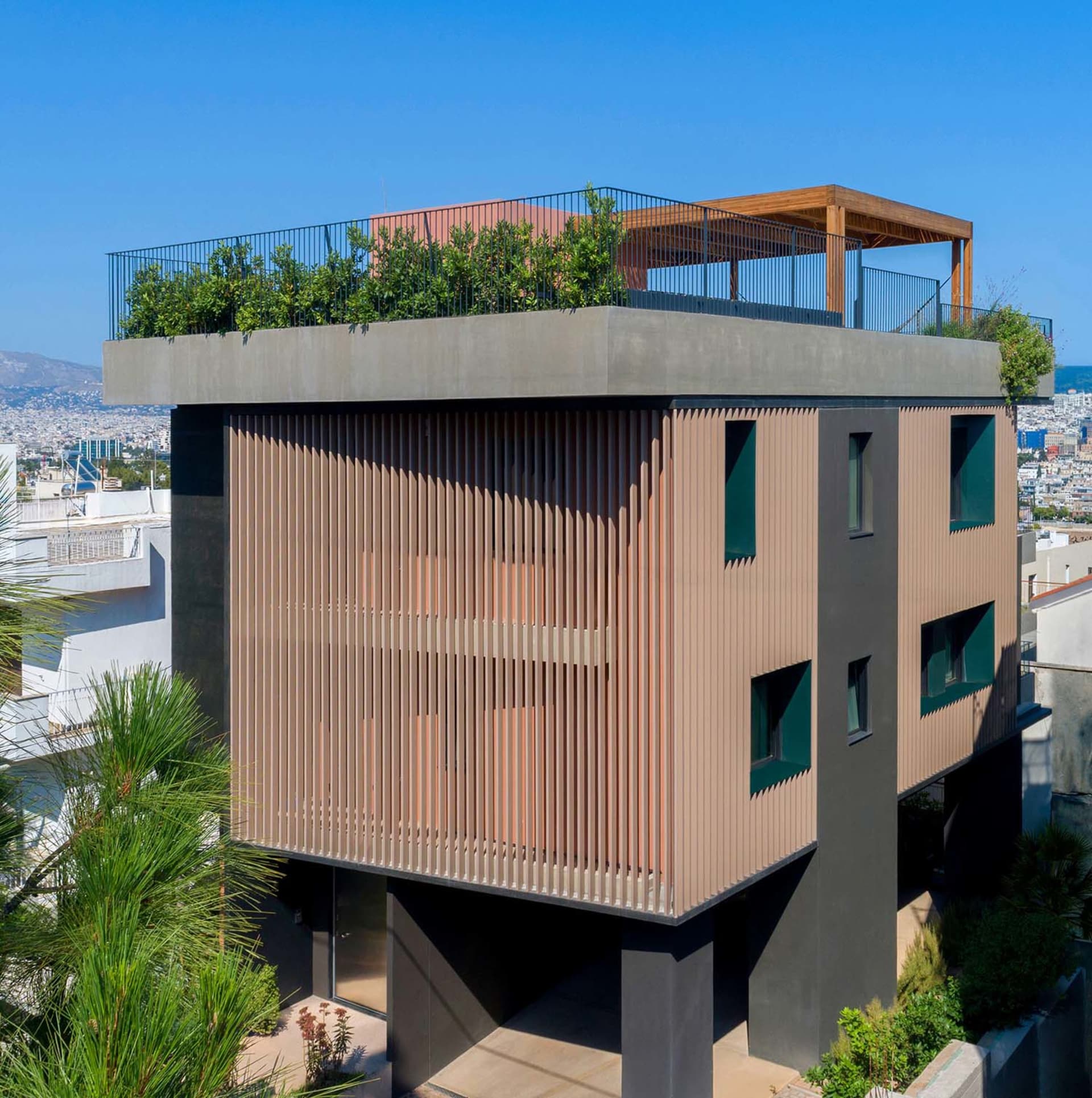 Modern building with corten steel and vertical wooden slats, featuring a lush rooftop terrace with wooden pergola against a clear blue sky and city view.