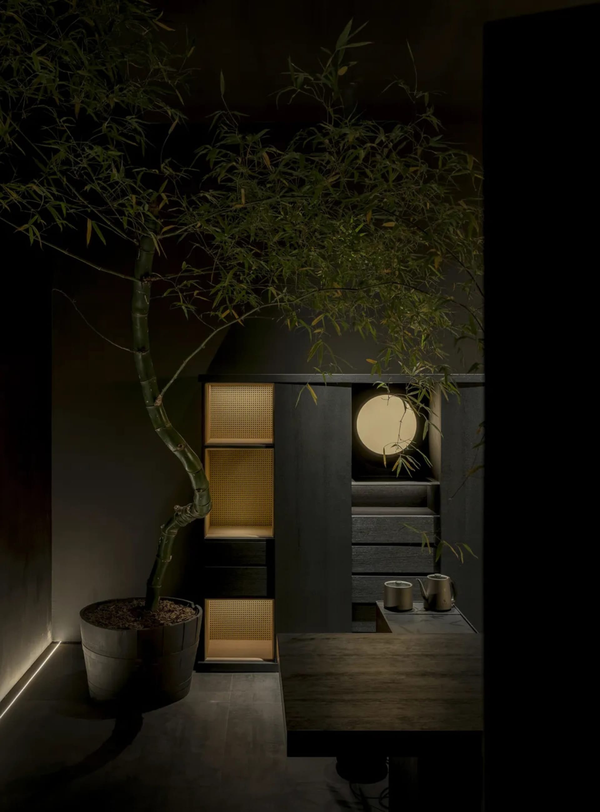 Modern minimalist tea room interior with a large potted bamboo tree, dark wood cabinetry with woven rattan panels, a glowing circular wall light, and a wooden table with tea set.