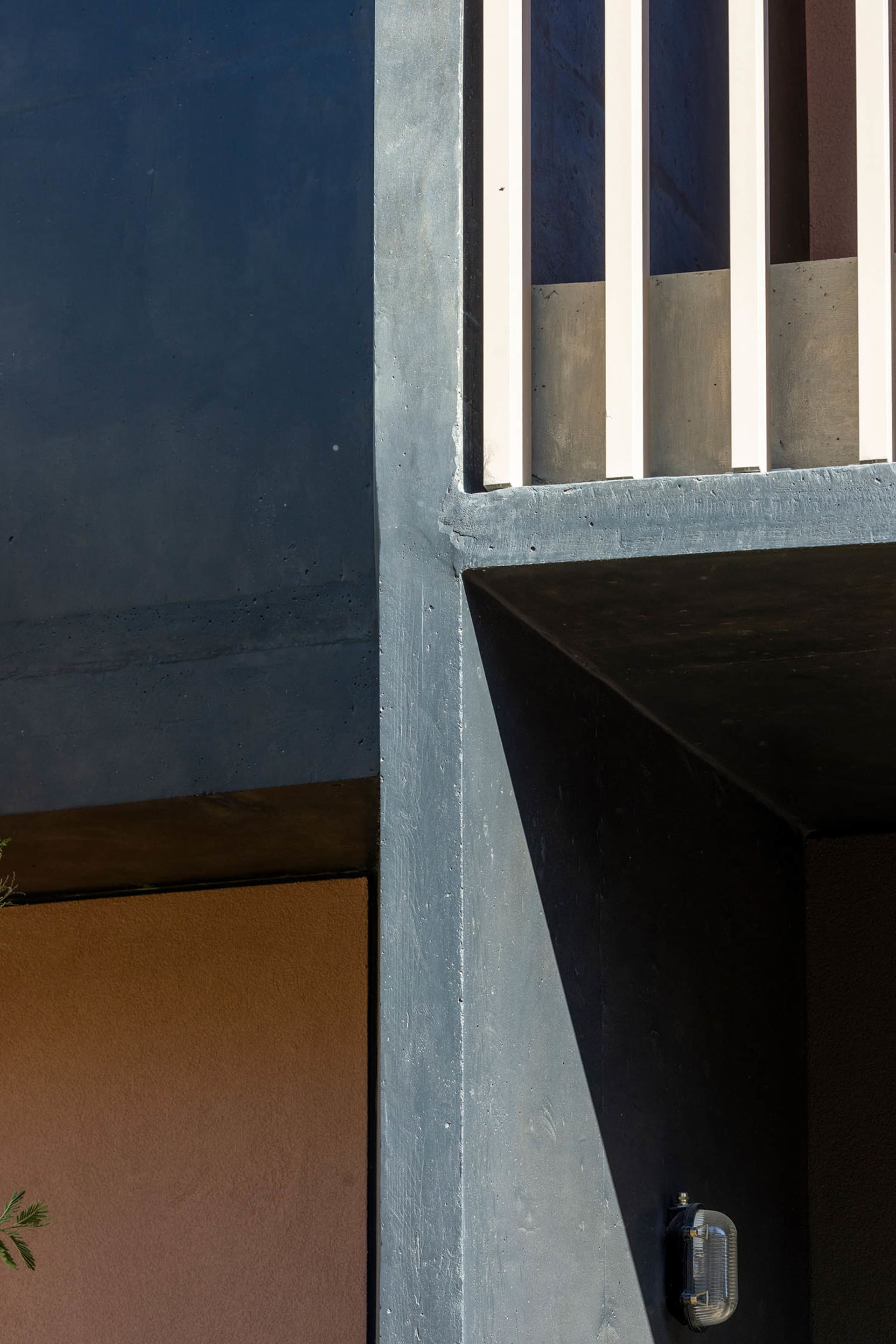 Close-up architectural detail of a raw concrete corner where deep blue, terracotta, and black painted surfaces meet under bright sunlight, creating sharp shadows.