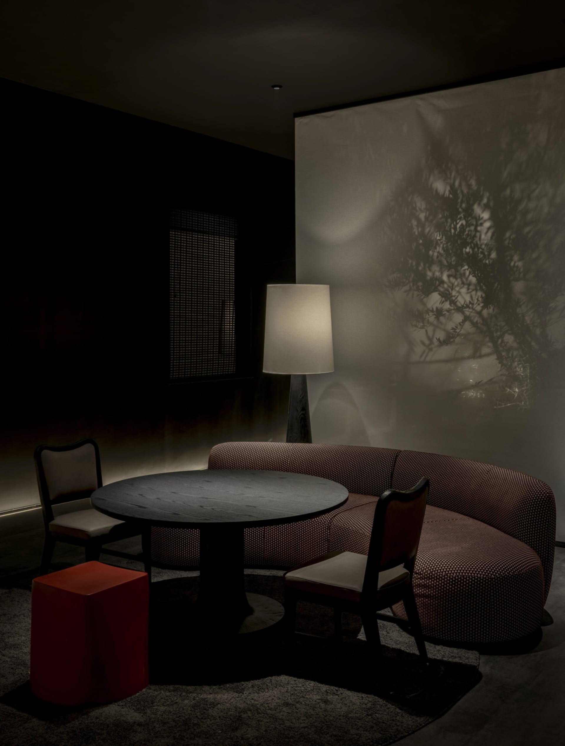 Modern moody dining area featuring a curved burgundy textured sofa, round dark wood table, matching chairs, a bright red cube stool, and dramatic tree shadow projection on the wall.