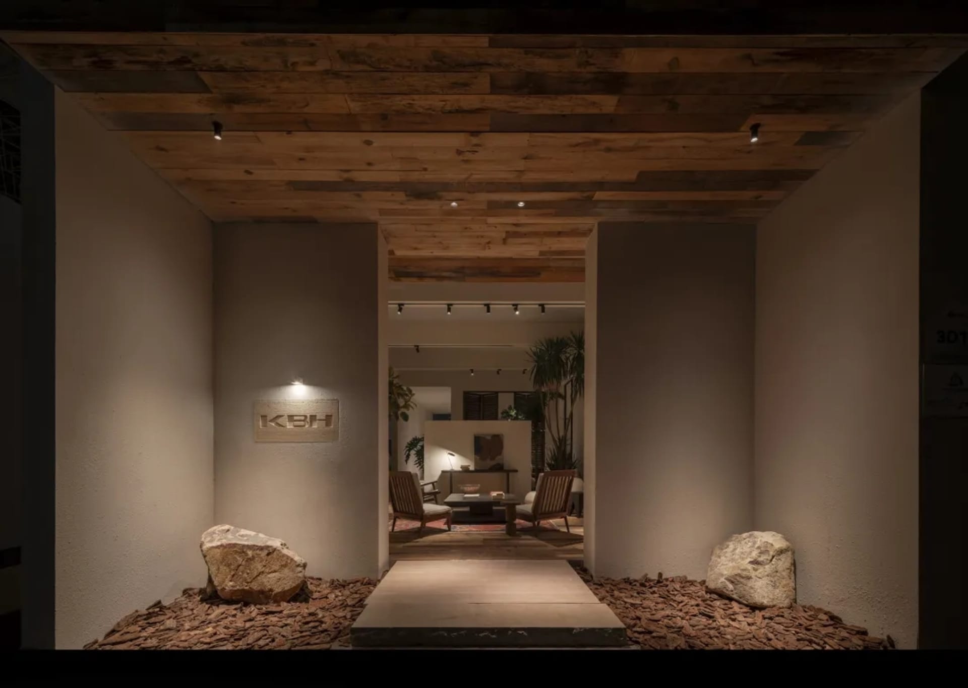 Modern entrance hallway featuring a reclaimed wood ceiling, textured beige walls, two large natural boulders on a bed of wood chips, and a view into a tropical-style living area beyond.