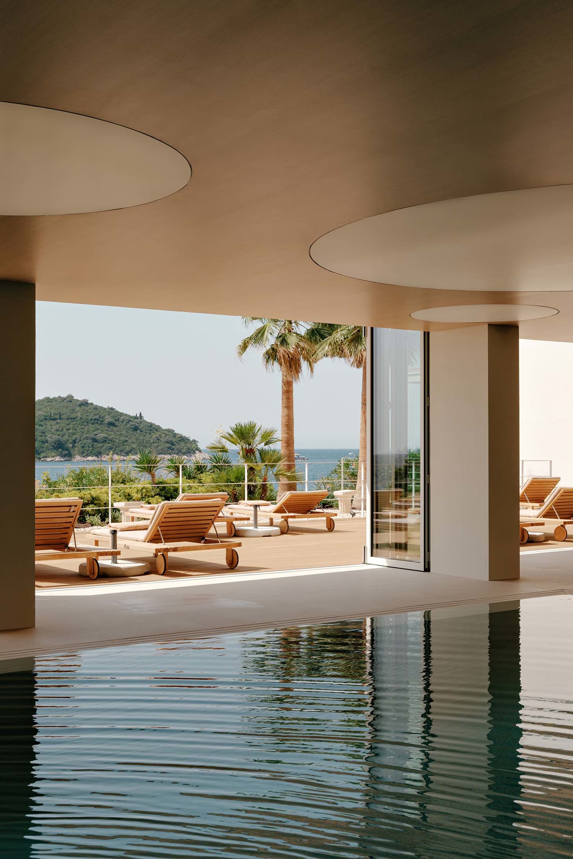 Modern infinity pool with wooden teak lounge chairs on a deck overlooking the Mediterranean sea and a green hillside under a clear sky.