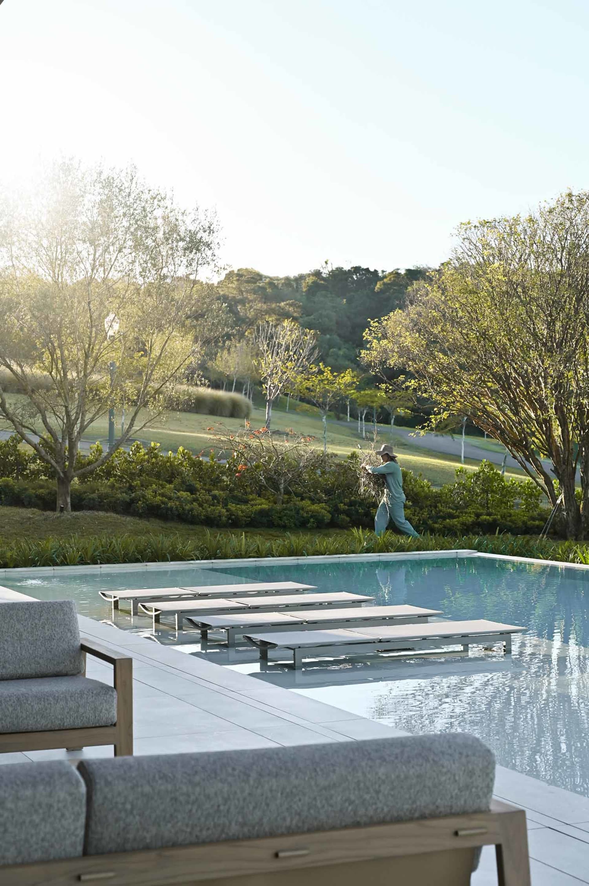 Modern infinity pool with teak and fabric lounge chairs on a stone deck, a person tending to plants in the background with forested hills beyond.