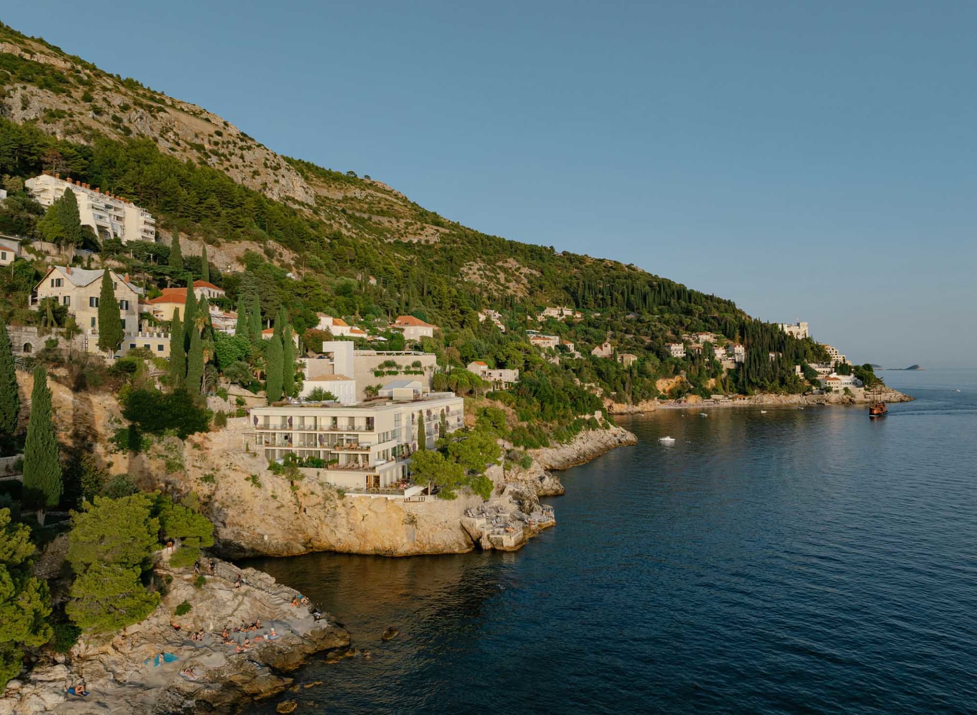 A modern white cliffside hotel built into rocky terraces overlooking the deep blue Adriatic Sea, with people sunbathing on the rocks below and green hills rising behind.