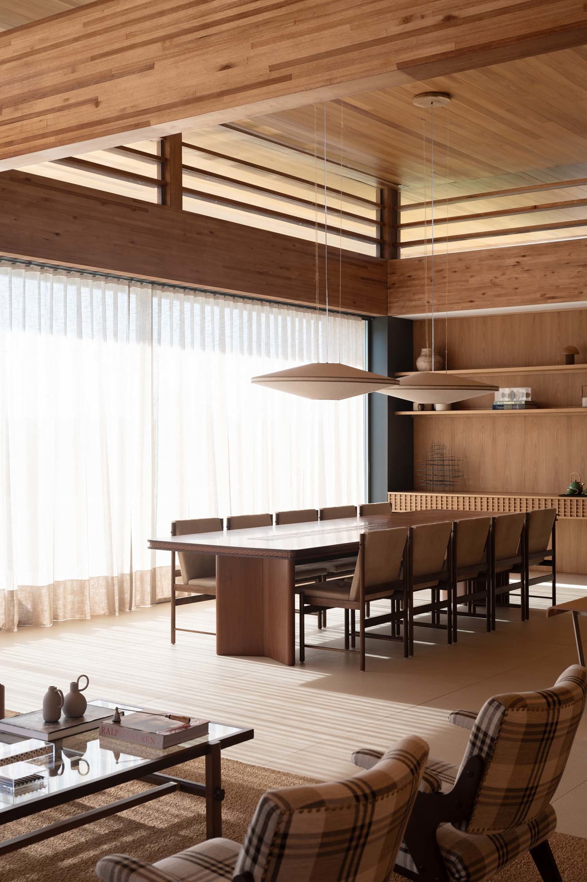 Modern dining room featuring a long walnut dining table with beige upholstered chairs, two large beige pendant lights, wooden ceiling beams, sheer curtains, and plaid armchairs in the foreground.