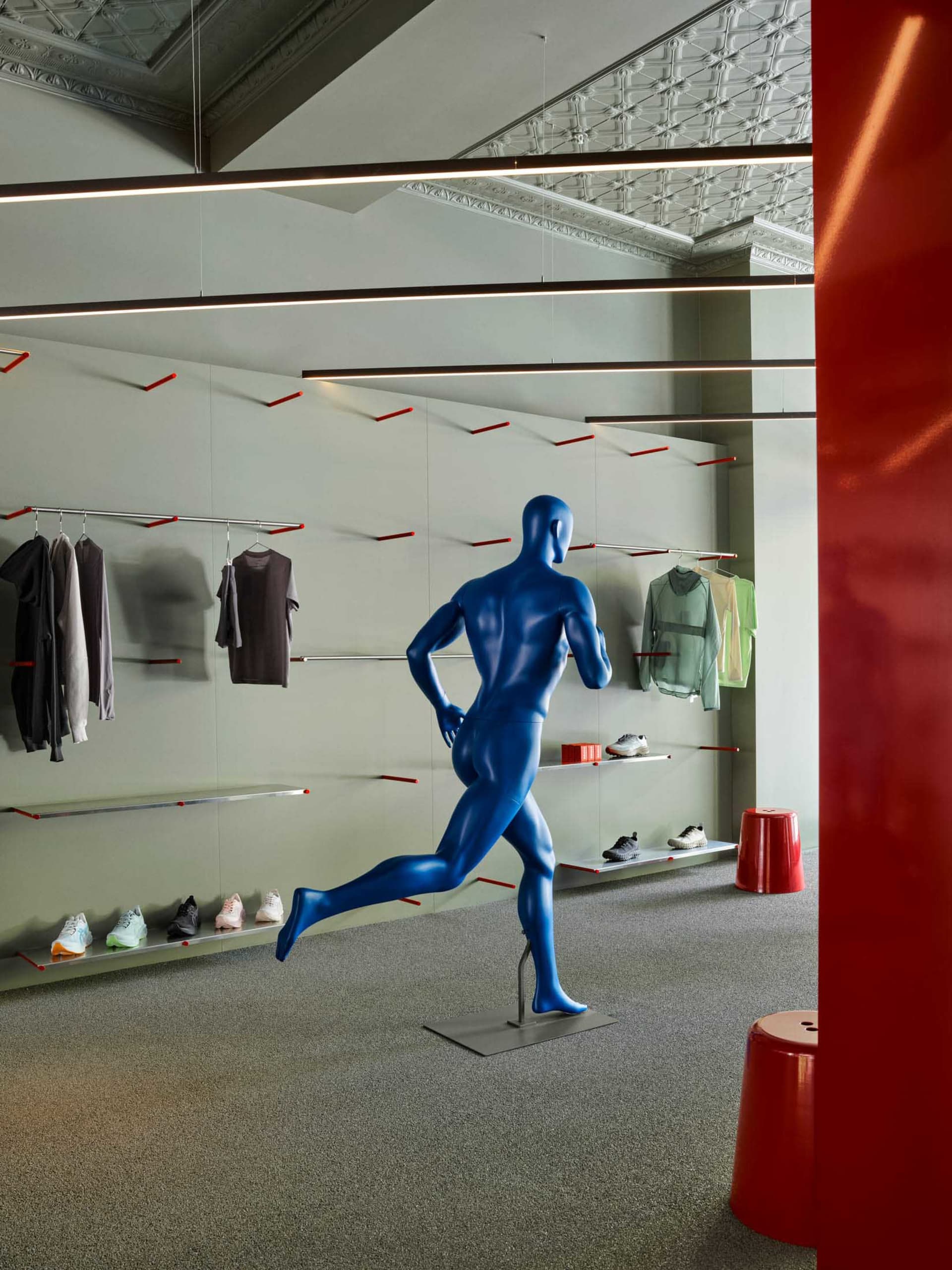 Interior view of a modern athletic apparel store featuring a glossy blue running mannequin, minimalist floating clothing racks with red accents, and linear LED lighting under an ornate ceiling.