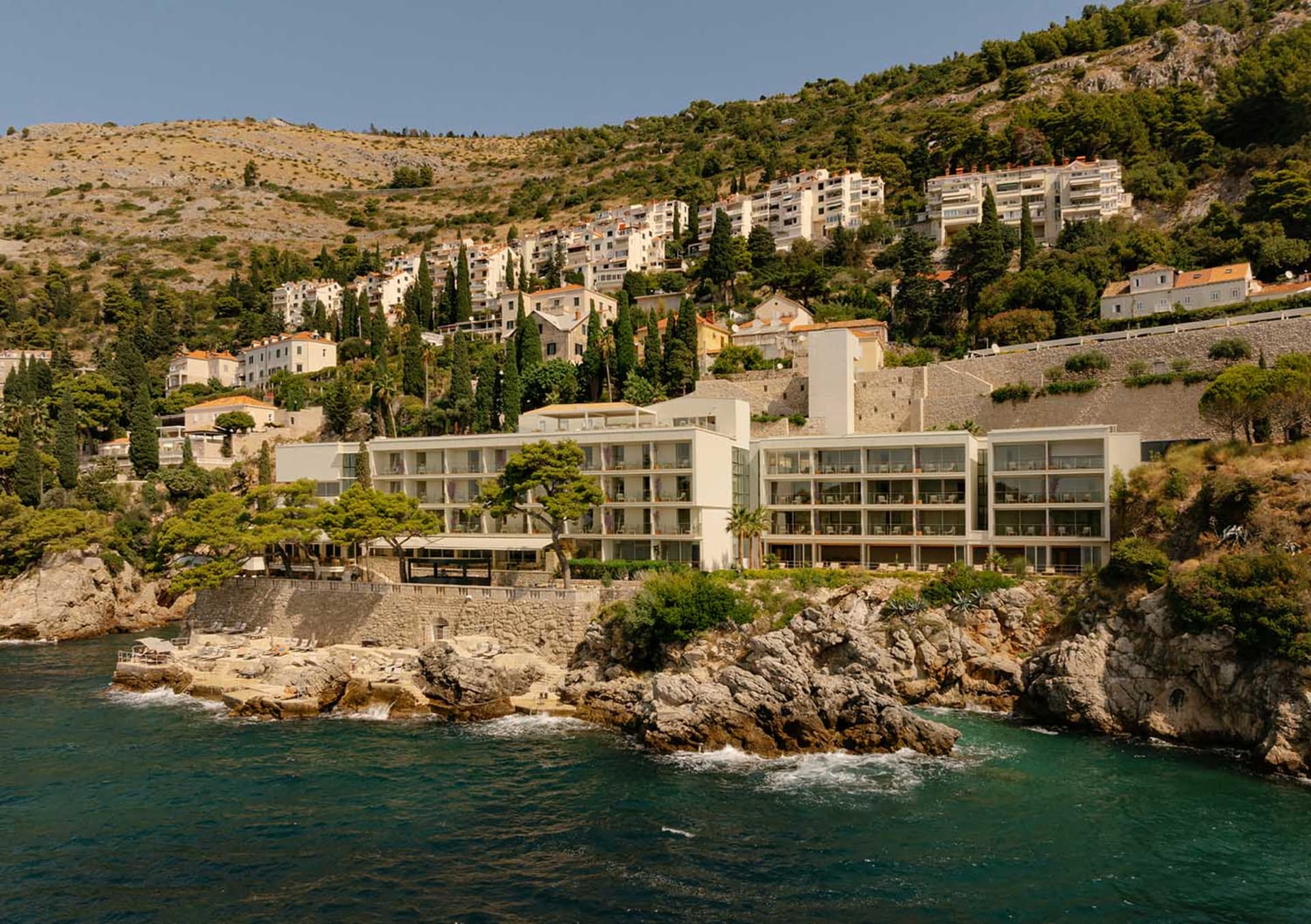 A contemporary seaside hotel built directly on a rocky coastline with turquoise water in the foreground and a hillside village rising behind it under a clear blue sky.