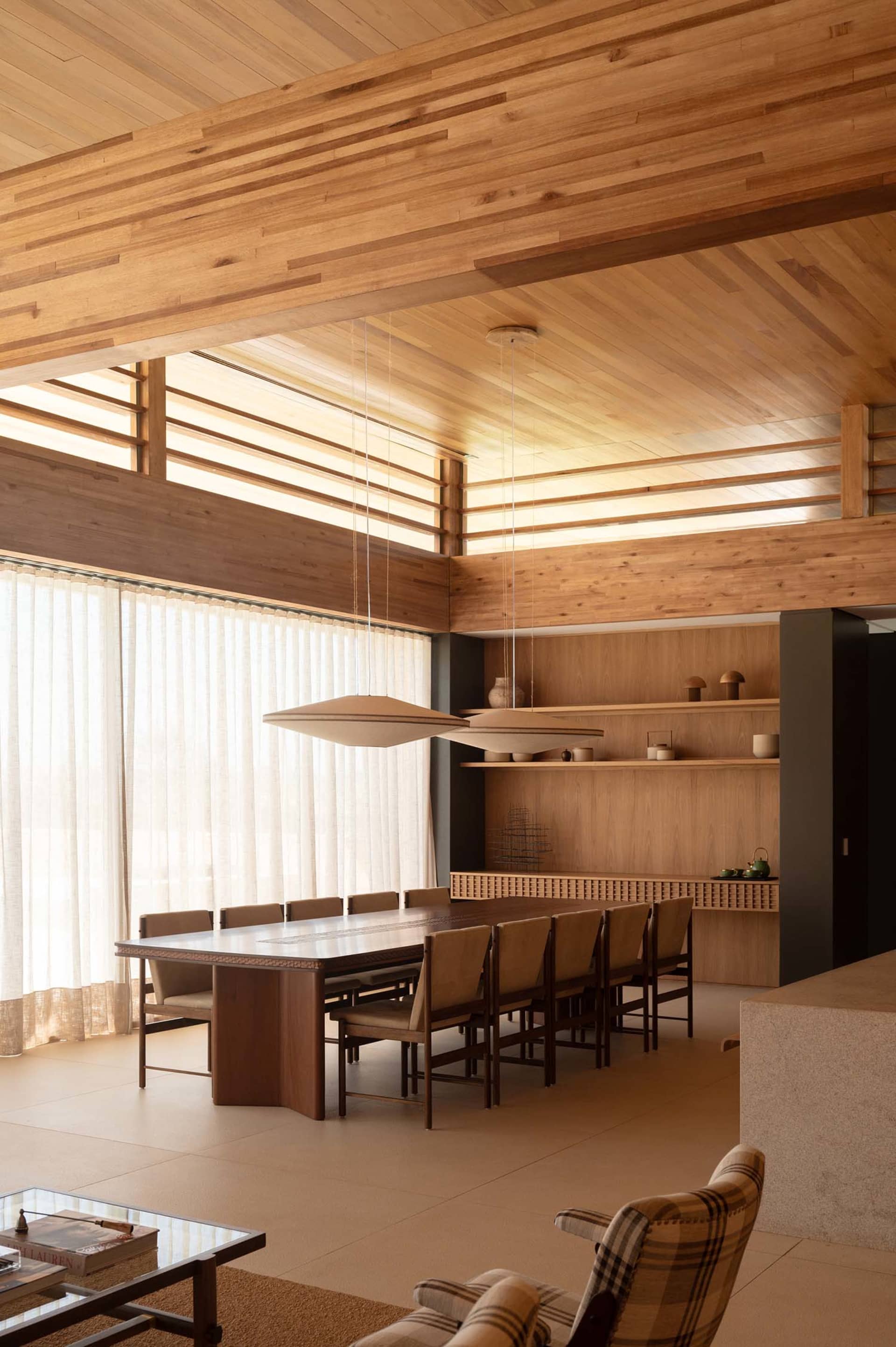 Modern dining room featuring an expansive wood-paneled ceiling, large walnut dining table with beige upholstered chairs, built-in wooden shelving, and a plaid armchair in the foreground.