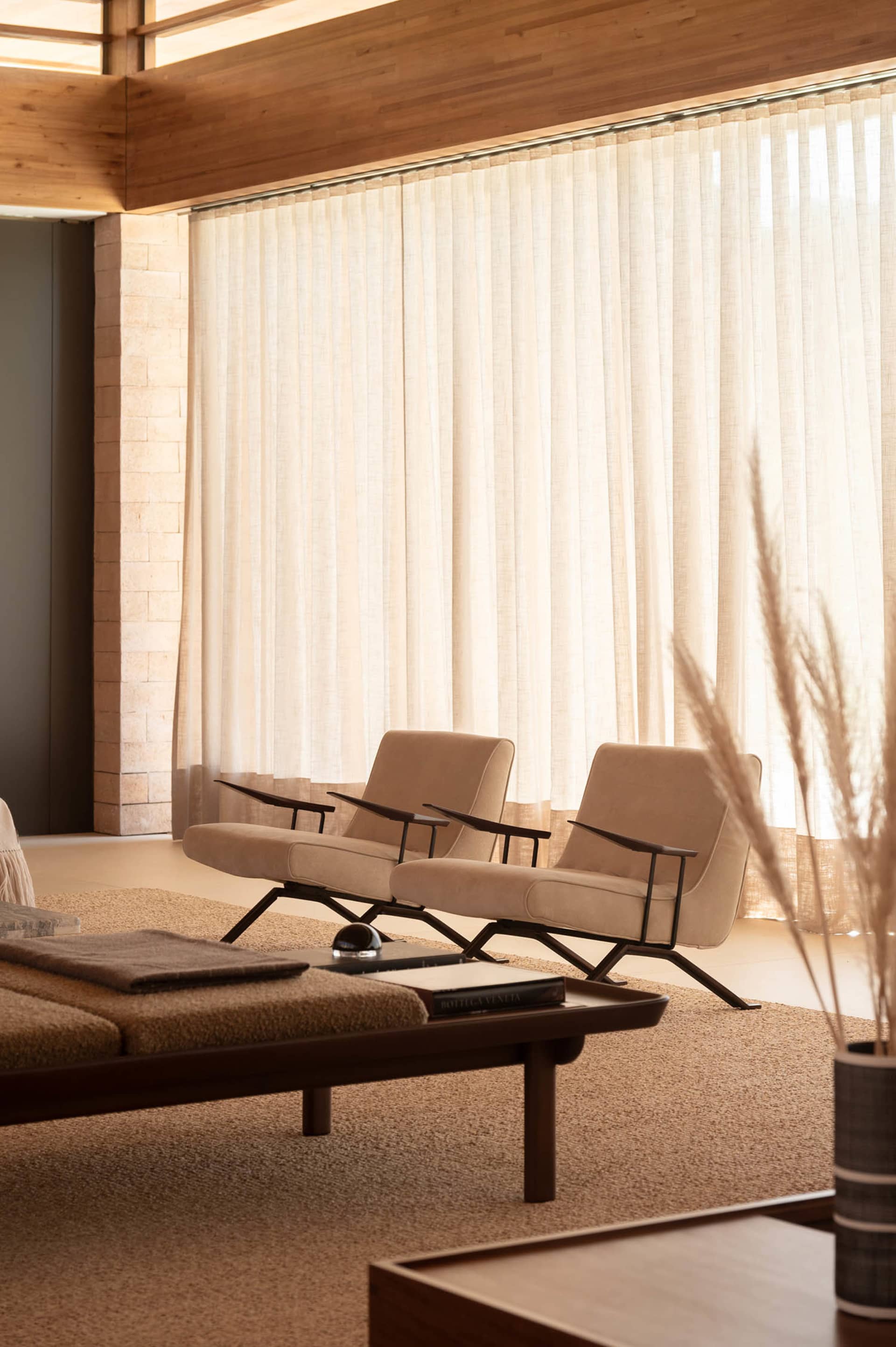 Modern minimalist living room featuring two beige upholstered lounge chairs with black metal frames, walnut coffee table, textured beige rug, stone wall, and sheer curtains with warm natural lighting.