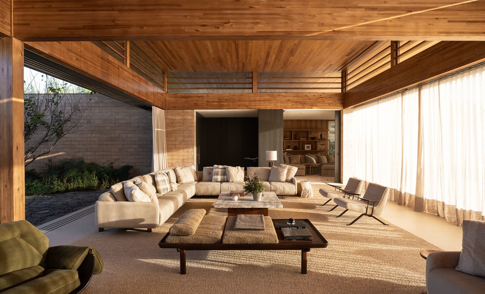 Spacious modern living room featuring a dramatic wood-paneled ceiling, large sectional sofa, natural fiber rugs, and warm sunlight streaming through sheer curtains.