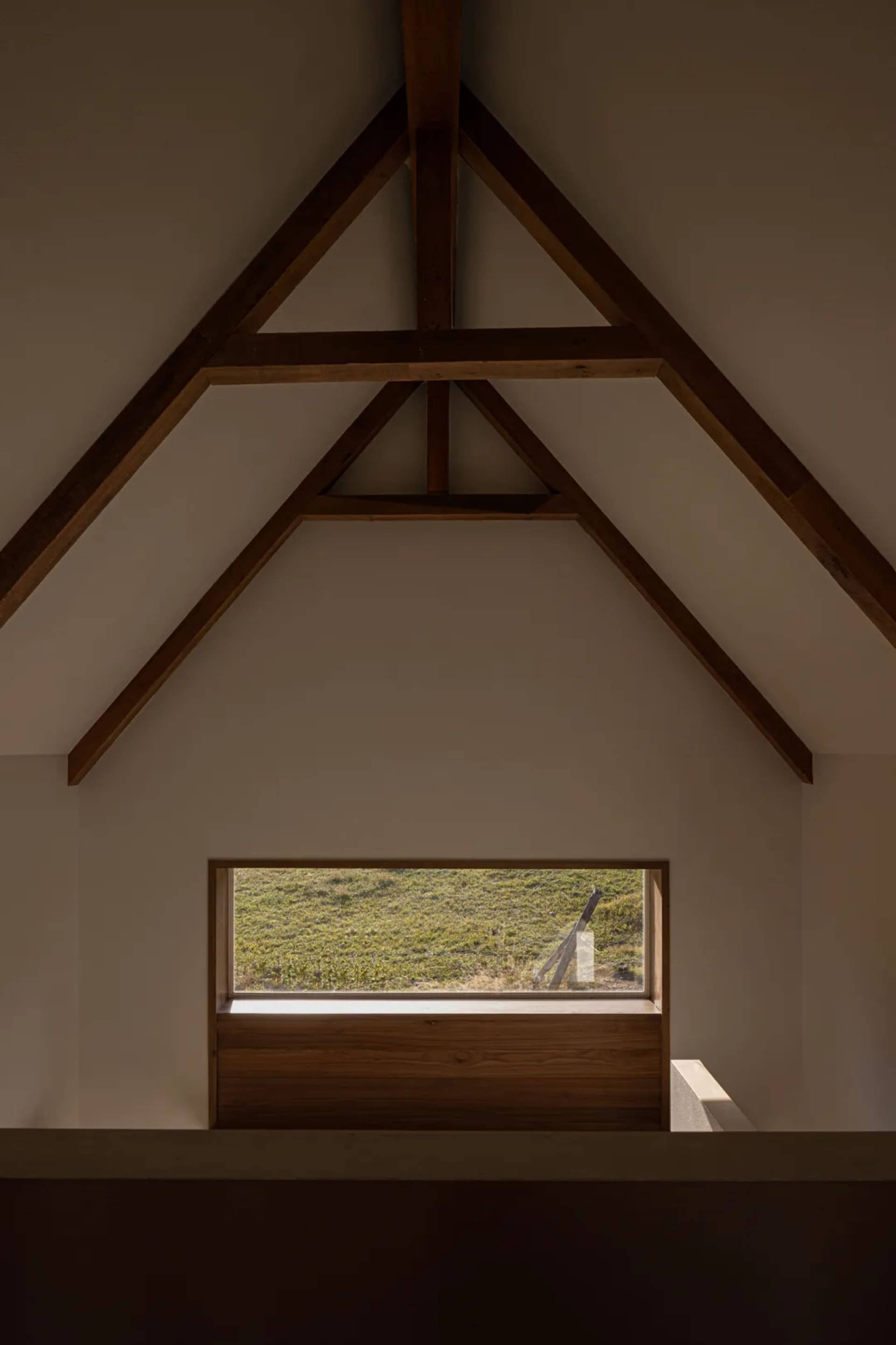 Interior view of a minimalist room featuring a dramatic exposed wooden gable ceiling, a built-in oak window seat, and a horizontal window overlooking a green grassy hill.